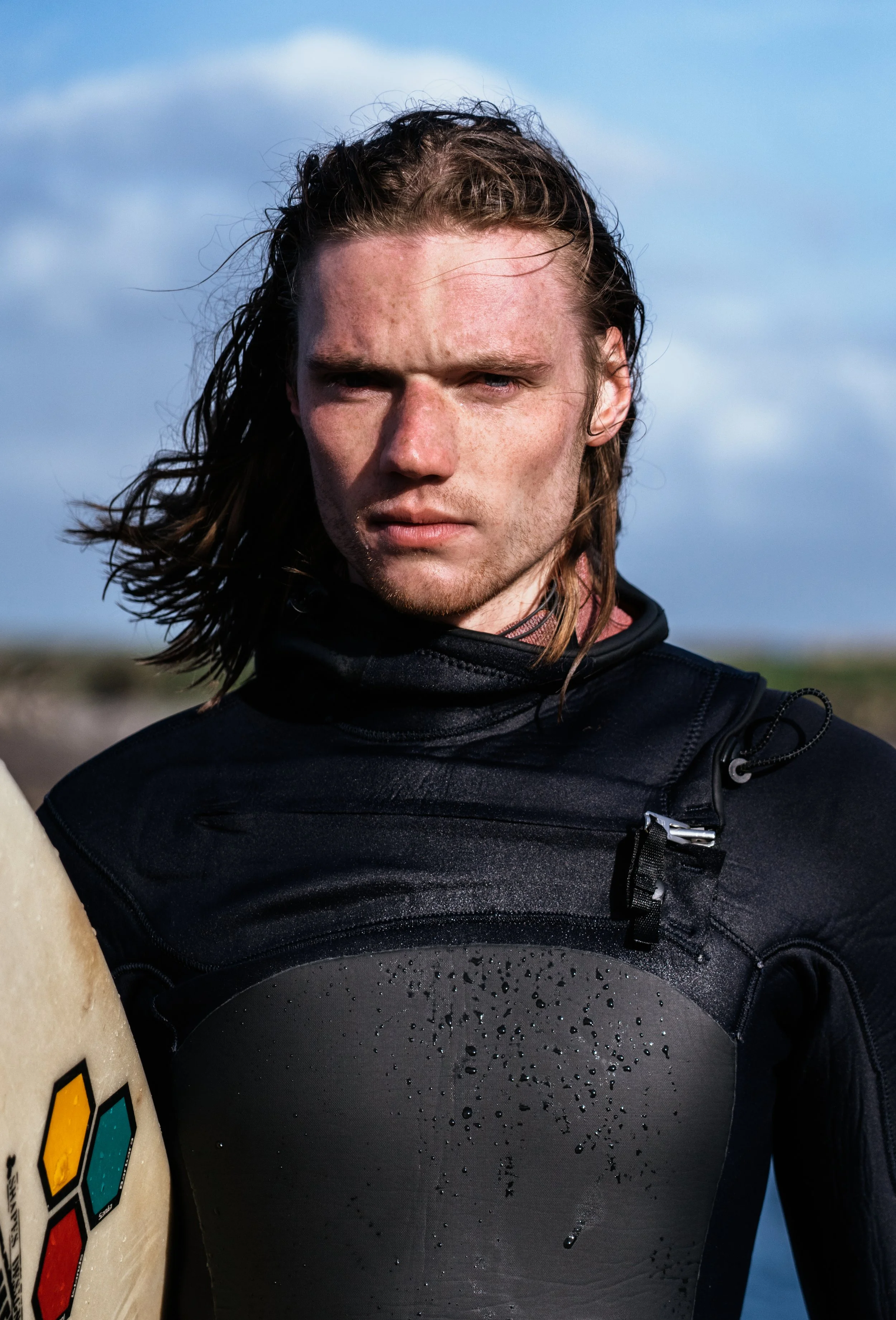 A young man with wet, long, brown hair outdoors, wearing a black wetsuit, holding a surfboard, with cloudy blue sky in the background.