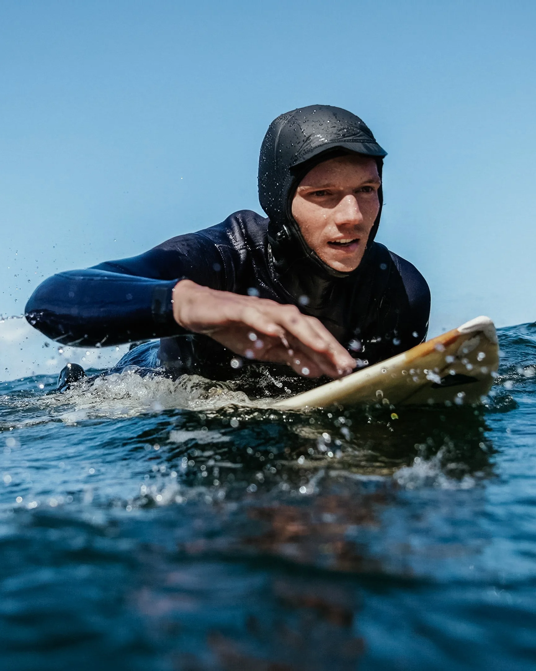 Male surfer in a wet suit and helmet paddling on a surfboard in the ocean under a clear blue sky.