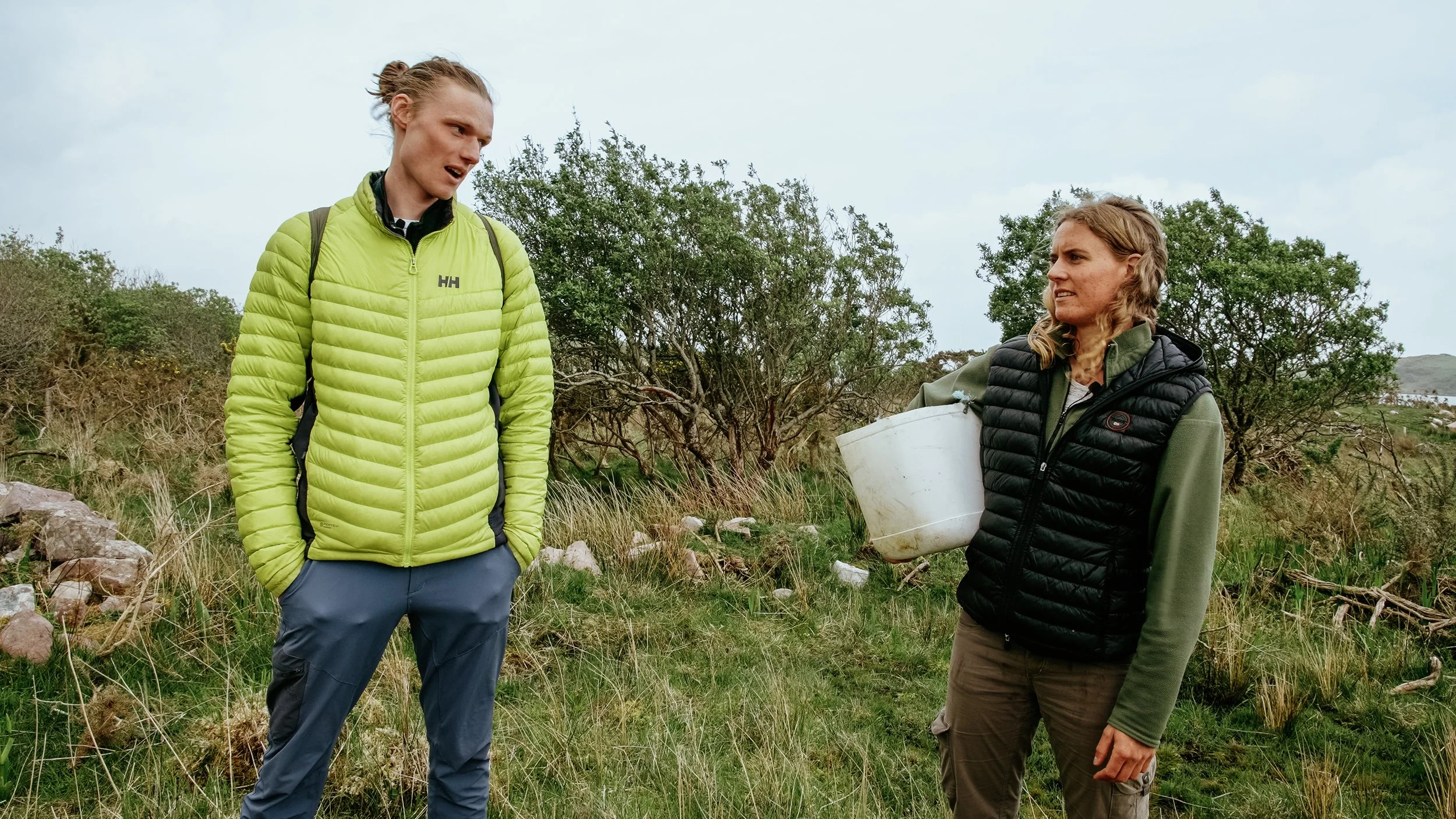 Two women outdoors in a natural setting, one holding a white bucket, engaged in conversation. One wearing a green jacket, the other a black vest.