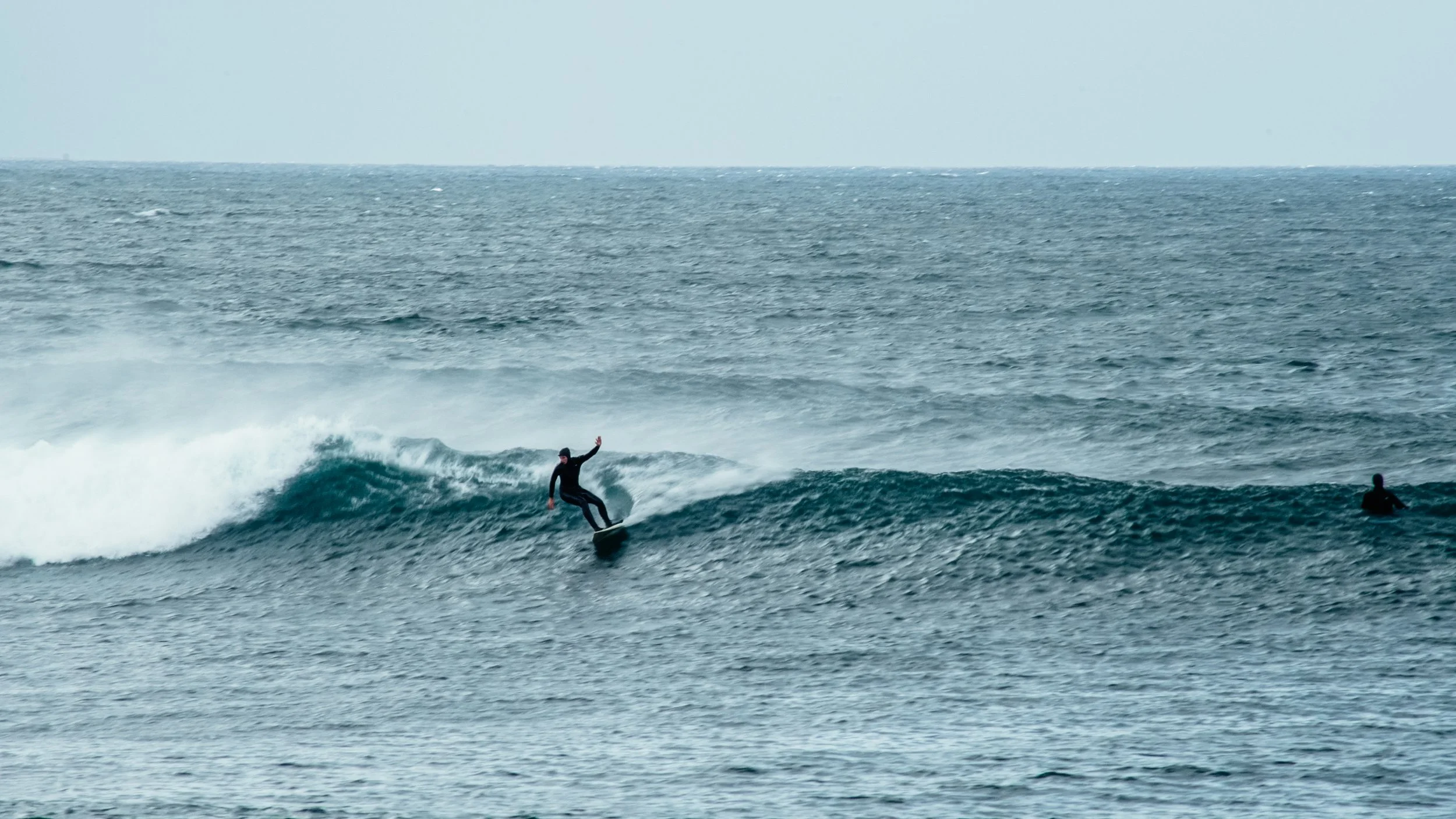 A person surfing on a wave in the ocean, wearing a black wetsuit, with another surfer in the water nearby.