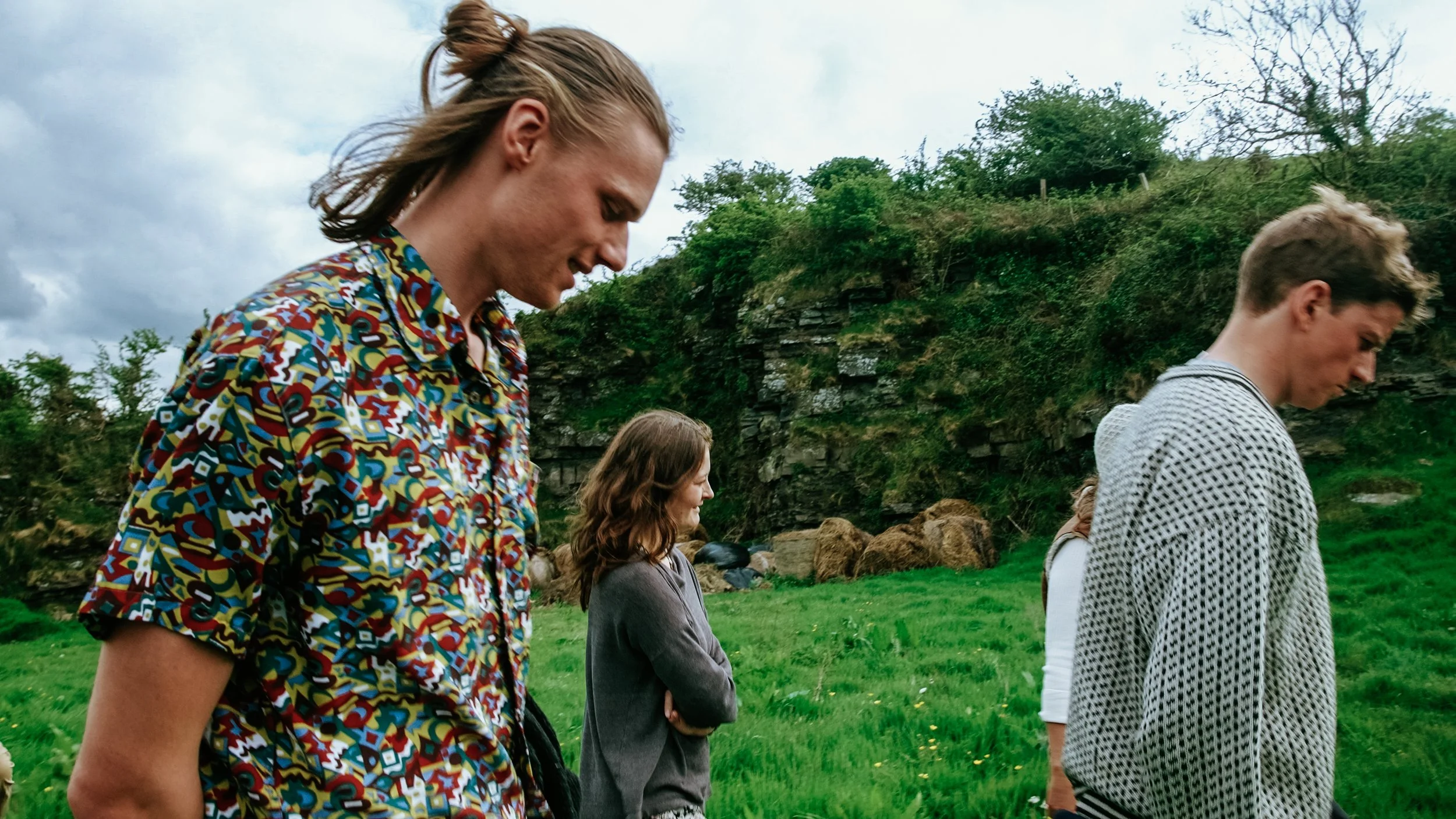 Four young people outdoors on a grassy area with rocky hills and trees in the background. They are standing in profile, looking downward, with their heads bowed.