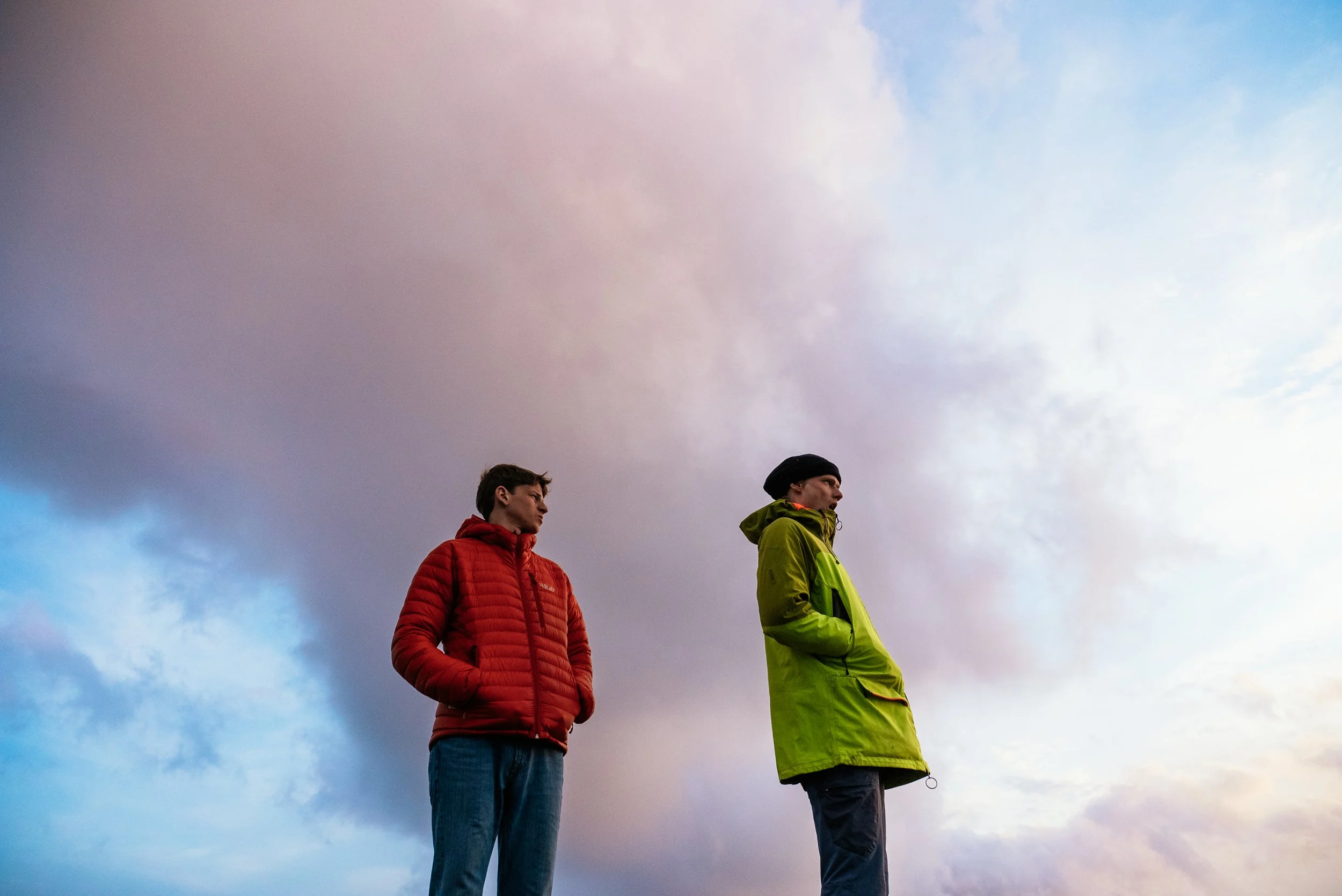 Two men standing outdoors under a cloudy sky, one in a red jacket and the other in a green jacket, both with hands in their pockets, looking to the side.