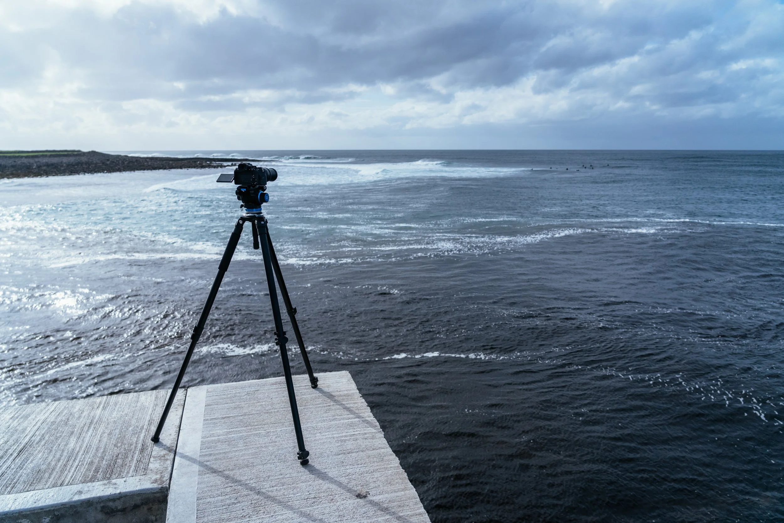 A camera on a tripod positioned on a concrete pier facing the ocean, with waves and a cloudy sky in the background.
