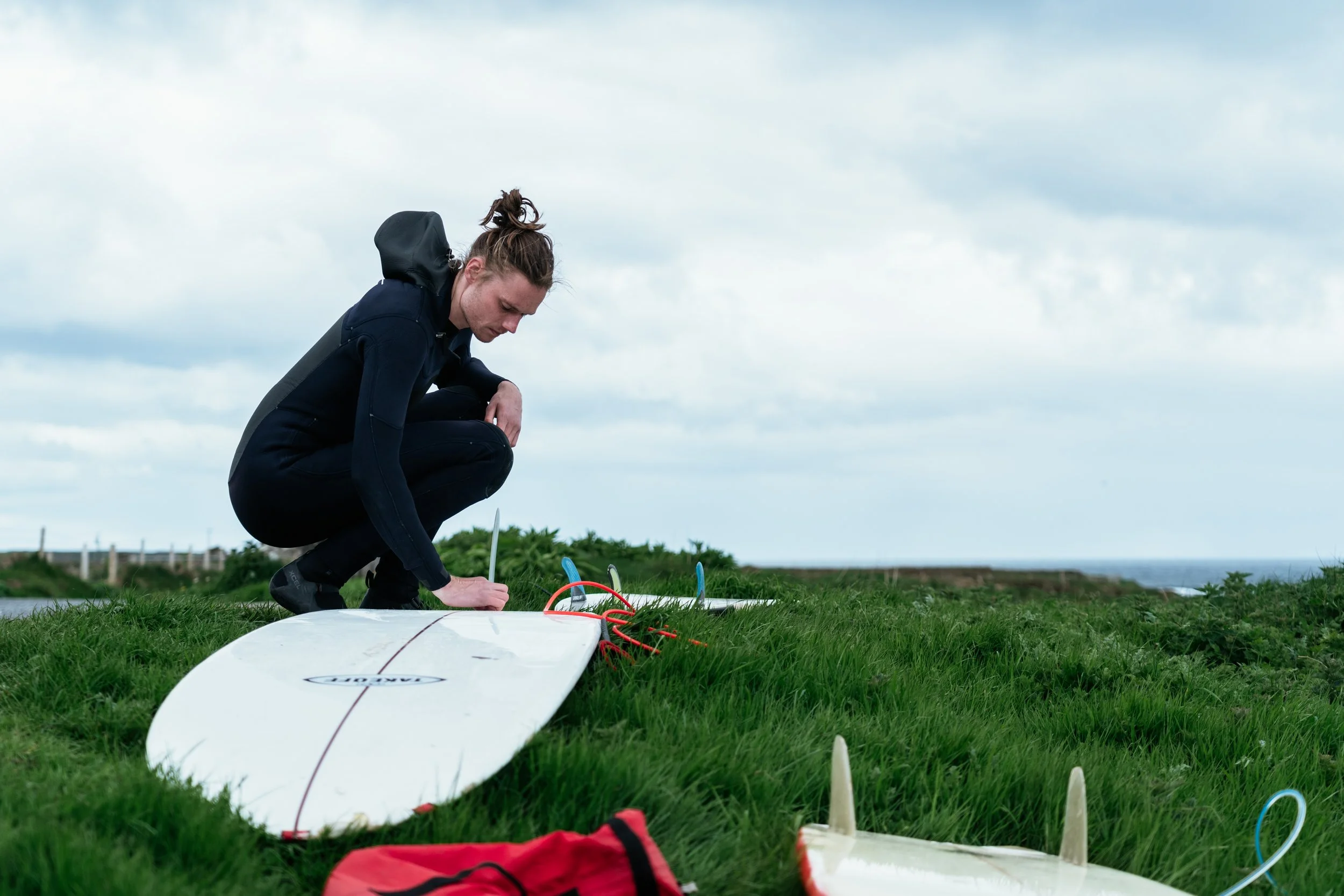 A person in black clothing preparing a surfboard on a grassy area outdoors under a cloudy sky.