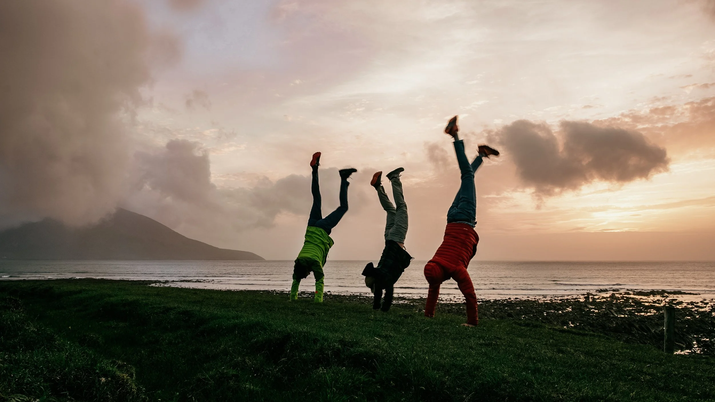 Three people do handstands on grass near the ocean at sunset, with mountains and clouds in the background.