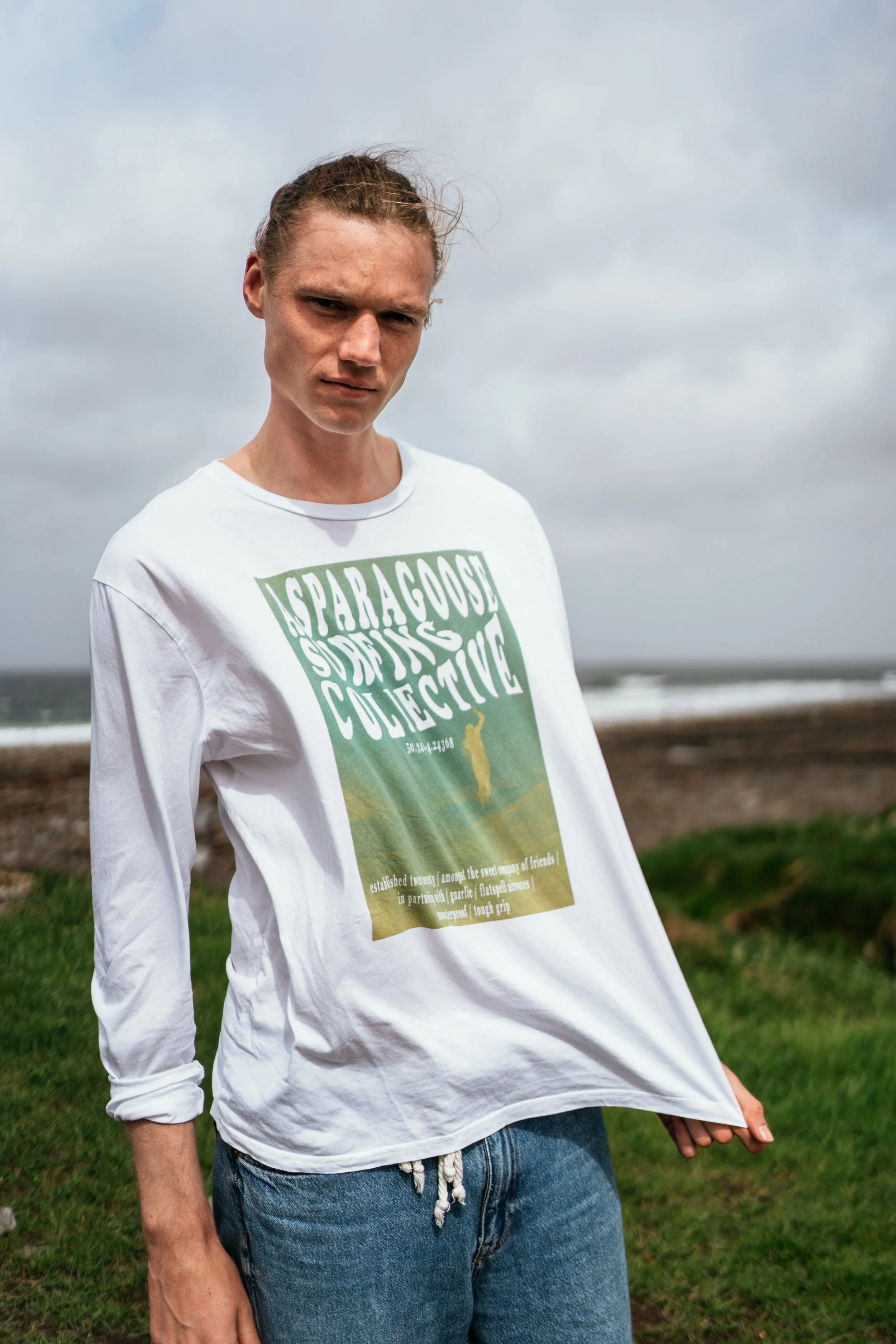 A young person with long hair in a bun, wearing a white long-sleeve t-shirt with graphic print, standing outdoors near the ocean on a cloudy day.