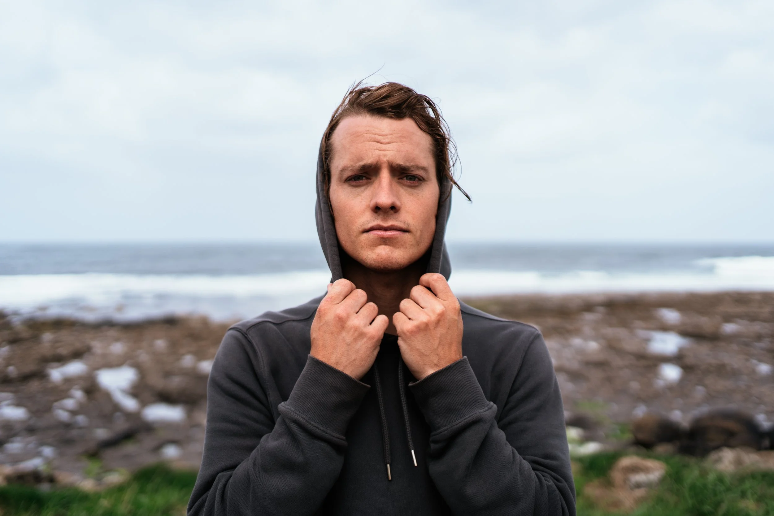 A young man with brown hair wearing a dark hoodie, holding the hood with both hands, standing outdoors near the shore with waves and rocks in the background, on a cloudy day.