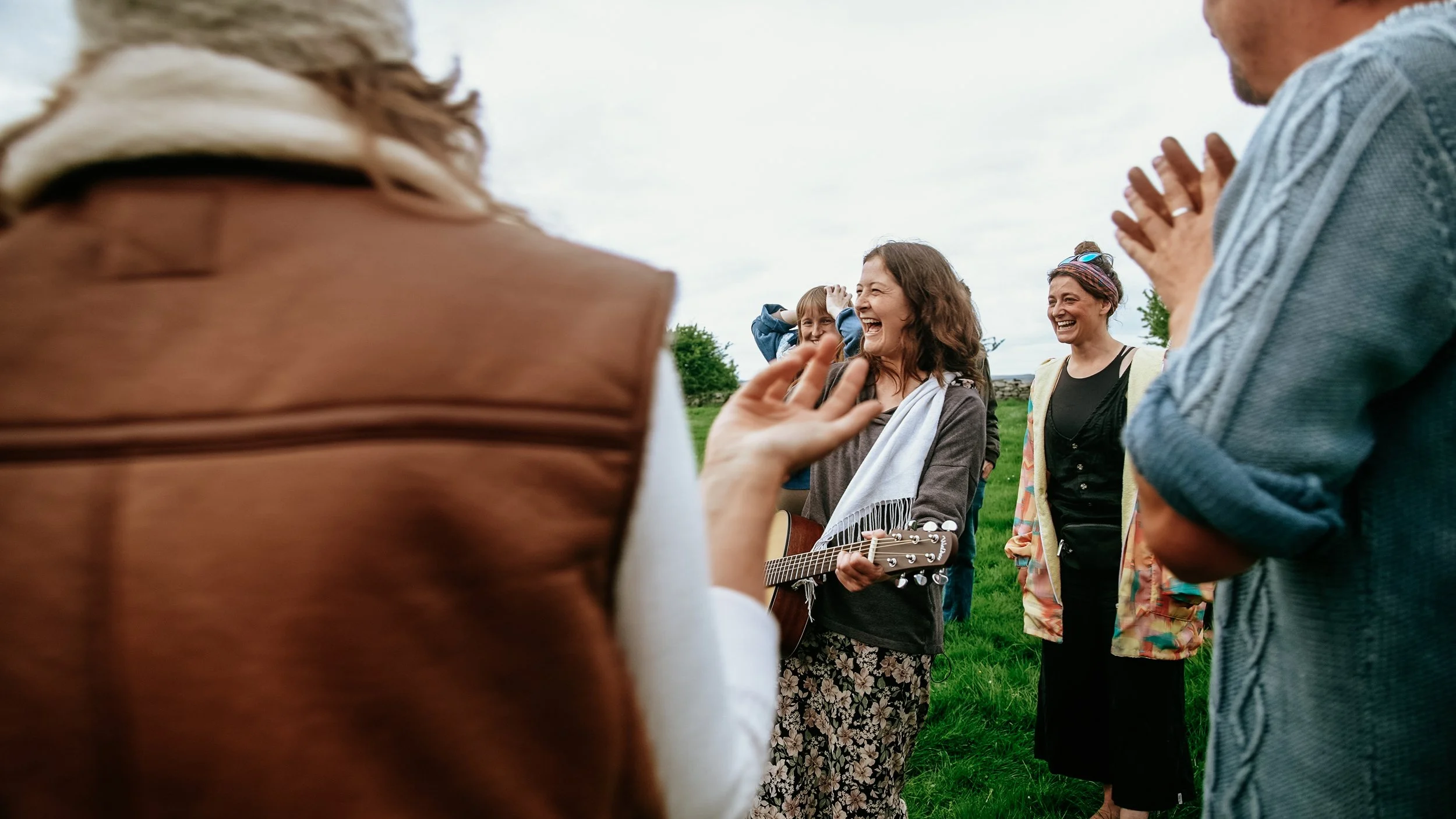 A group of people outdoors, with one woman playing guitar, smiling and laughing among friends.