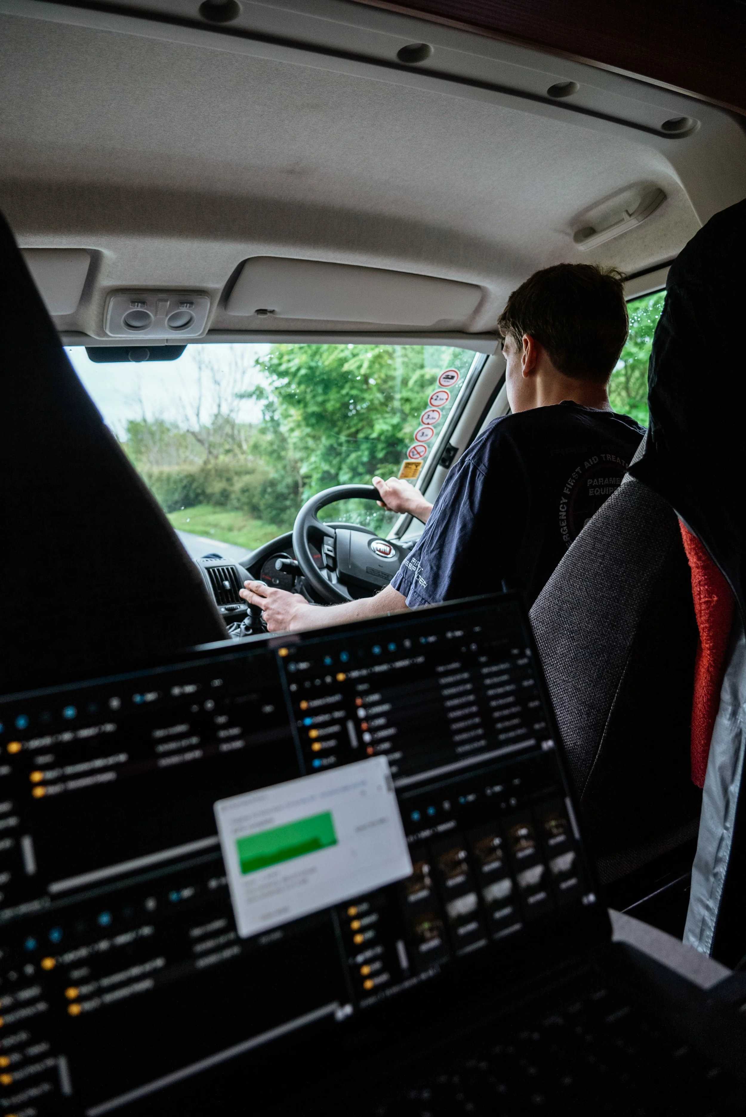 Person driving a vehicle with greenery outside the window, seen from the back of the vehicle with a laptop in the foreground.