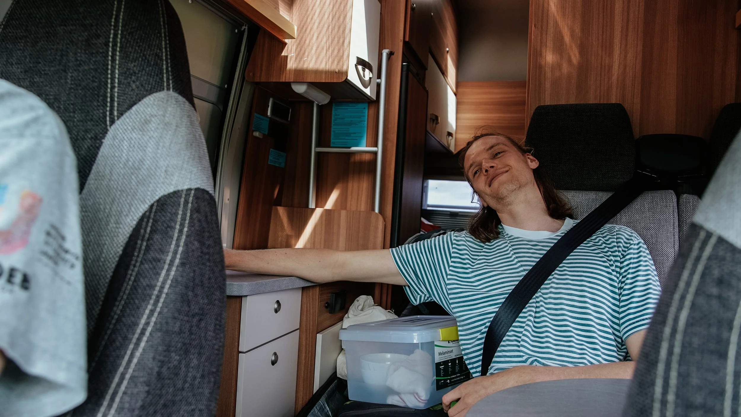 A man with long hair and a striped shirt sitting in a vehicle, smiling, with his arm extended to reach a surface. The vehicle has wood-paneled walls and compartments, and the man is secured with a seatbelt.
