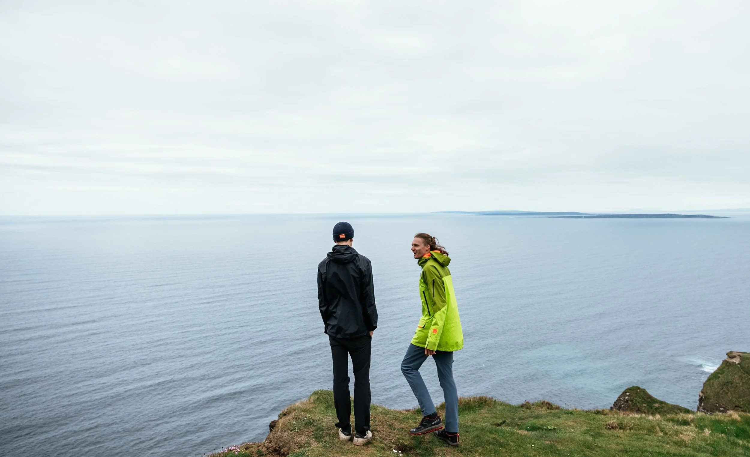 Two people stand on a grassy cliff overlooking a calm ocean, talking to each other.
