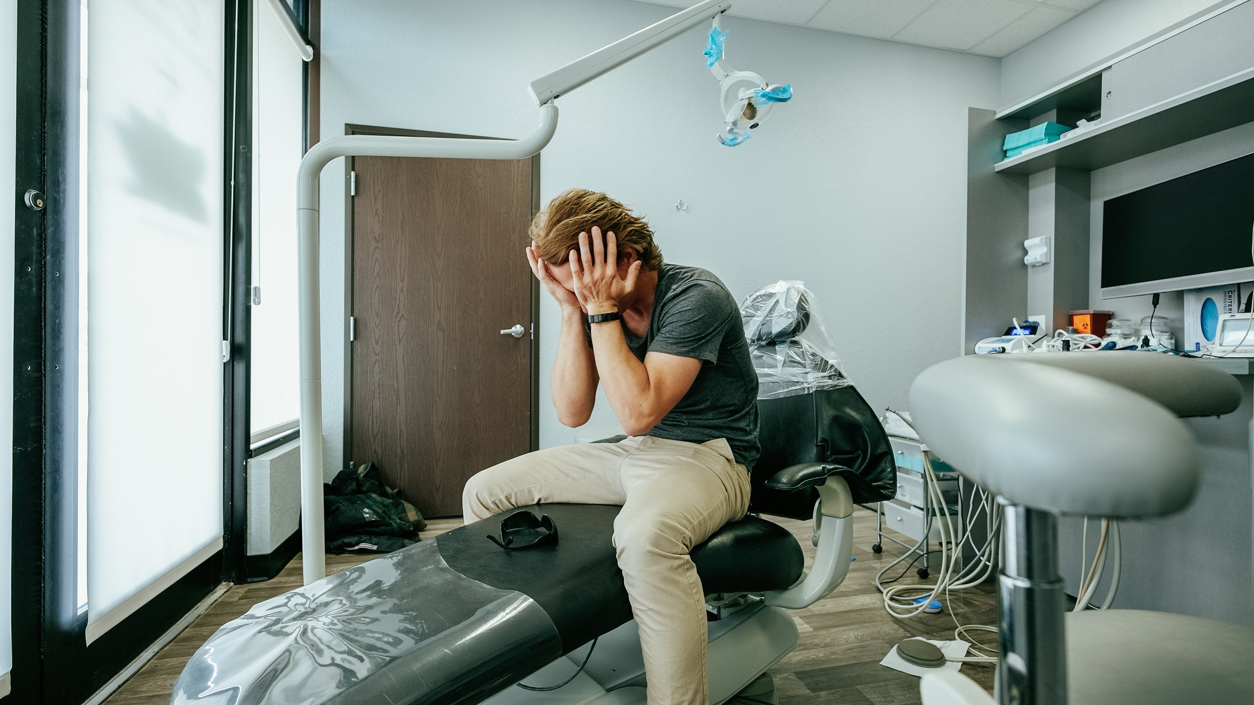 A man sitting on a dental examination chair with his hands covering his face, appearing distressed or overwhelmed in a dental office with medical equipment and supplies.