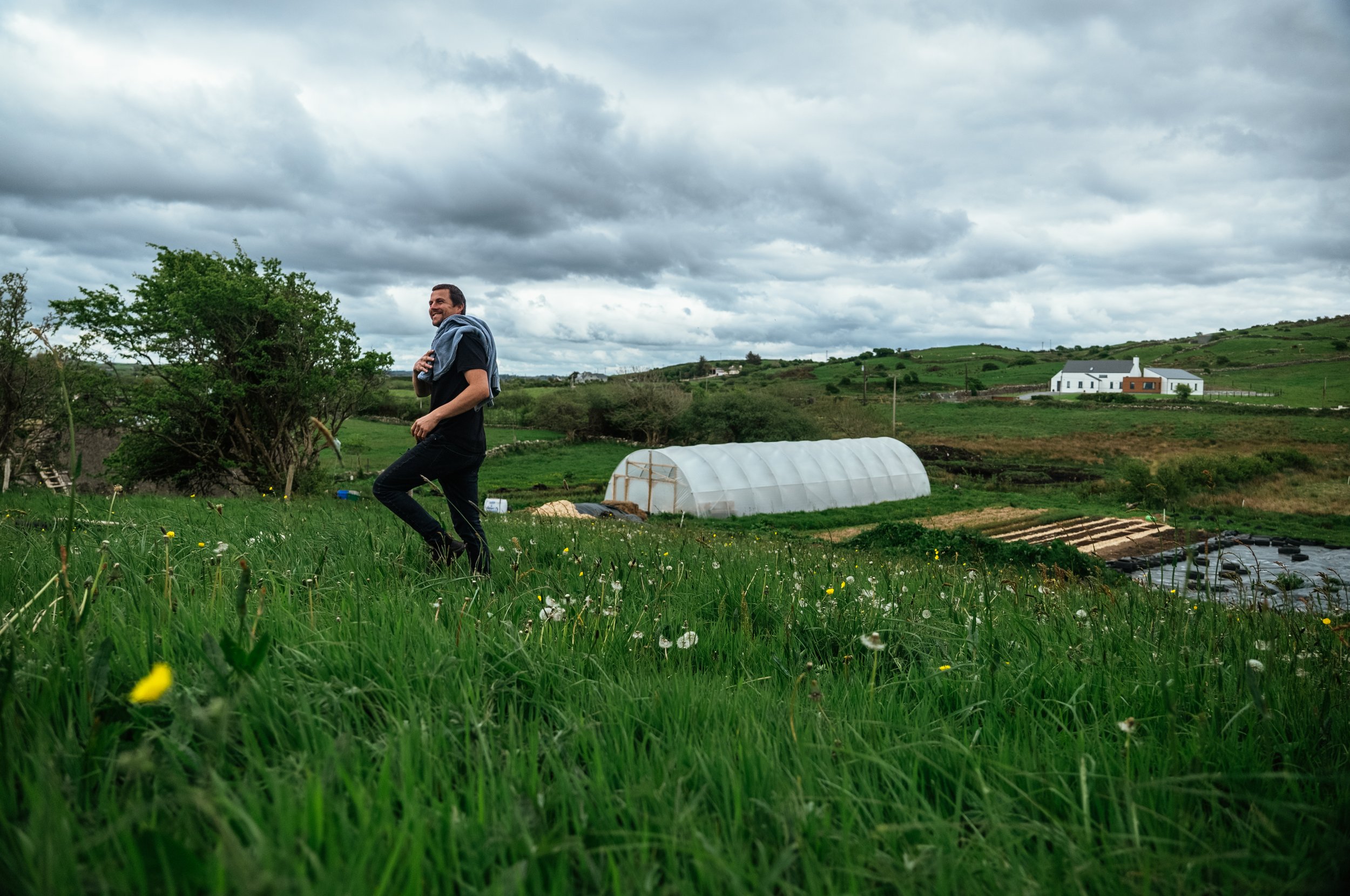 A man running through a grassy field with dandelions, holding a jacket on his shoulder, in a rural landscape with greenhouses and houses under a cloudy sky.