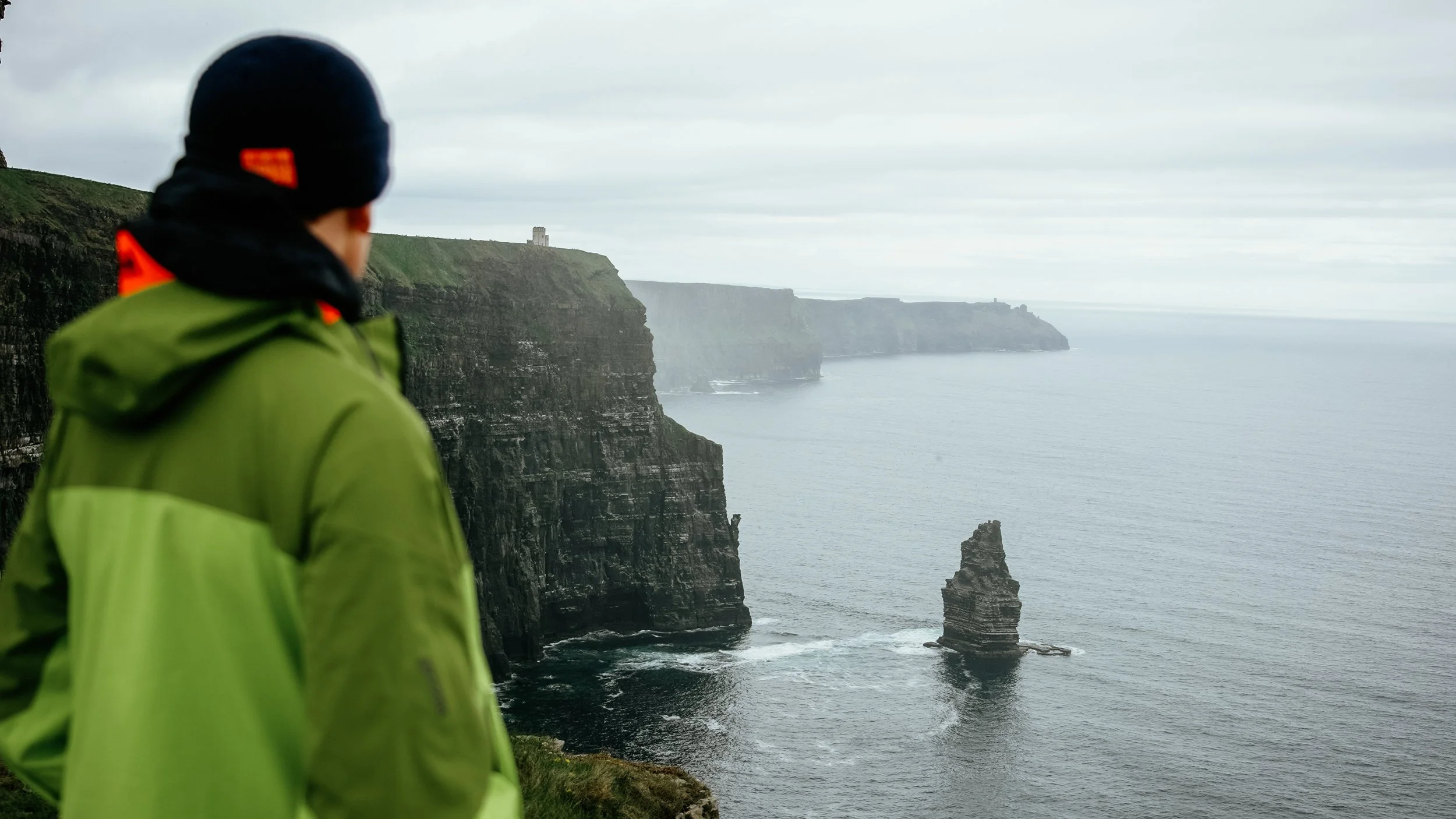 Person wearing a green jacket and black hat overlooking the Cliffs of Moher in Ireland, with a sea stack and cloudy sky in the background.