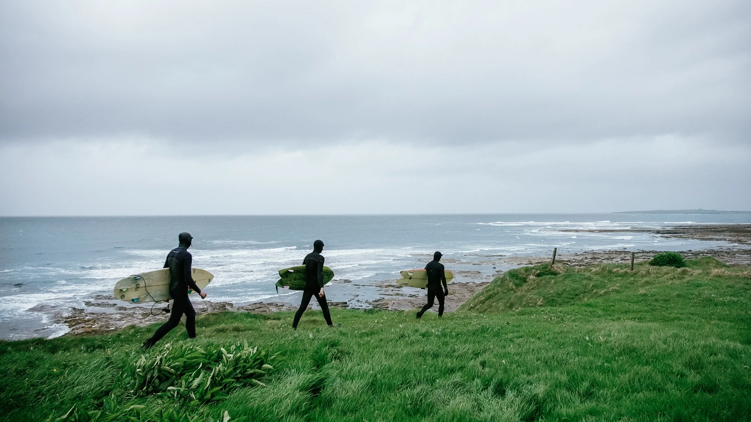 Three surfers dressed in black wetsuits walking towards the ocean carrying surfboards on a cloudy day.