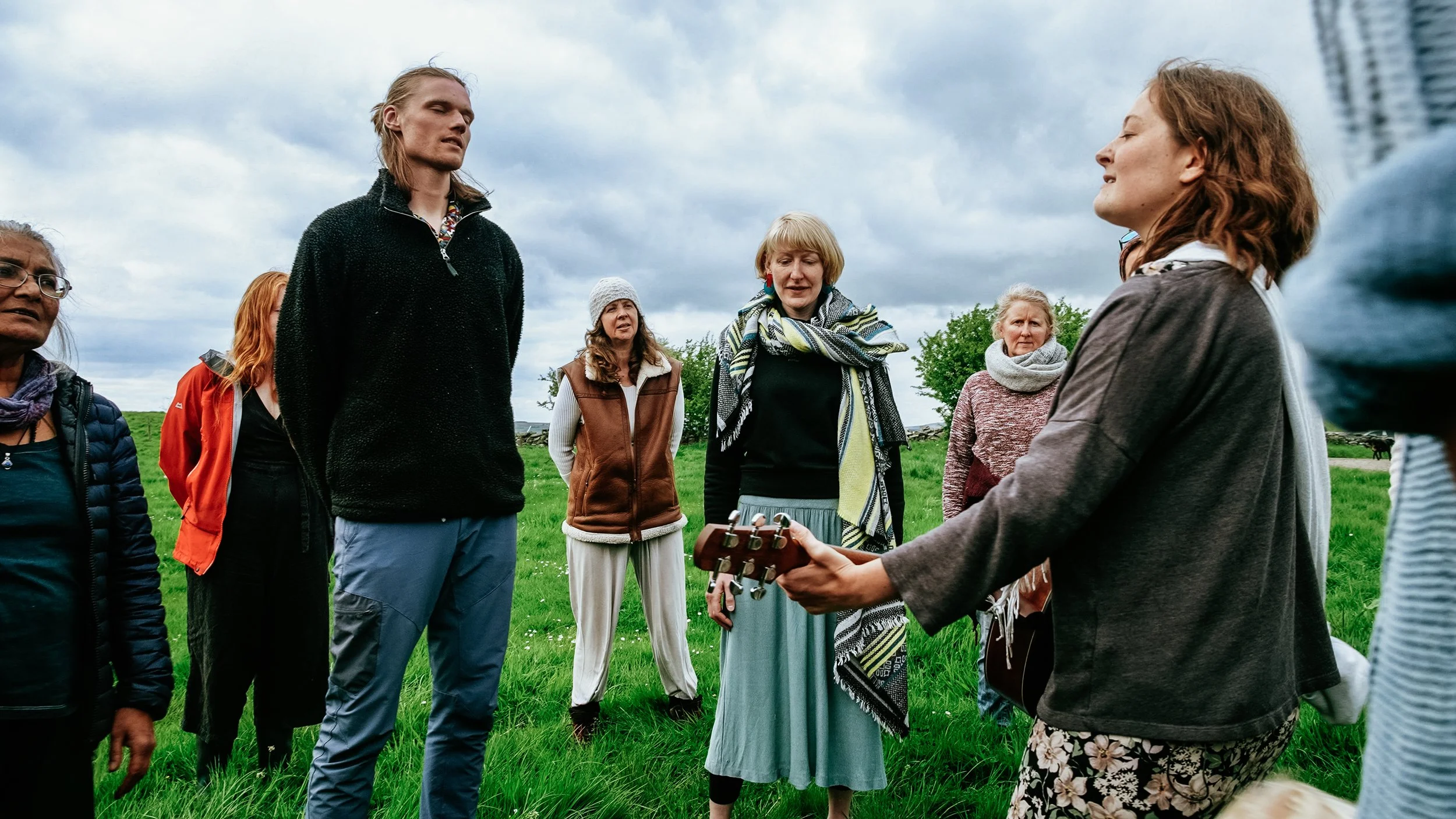 Group of people standing outdoors on a grassy field, some with their eyes closed, listening to a woman playing guitar, with cloudy skies overhead.