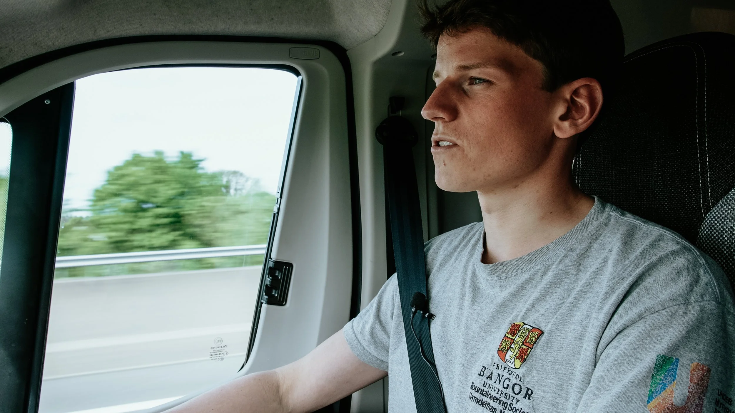 A man with short brown hair and fair skin is sitting in the driver's seat of a vehicle, holding the steering wheel. He is wearing a gray T-shirt with university and coat of arms logos on the front, and a seatbelt. Outside the window, a green, blurred