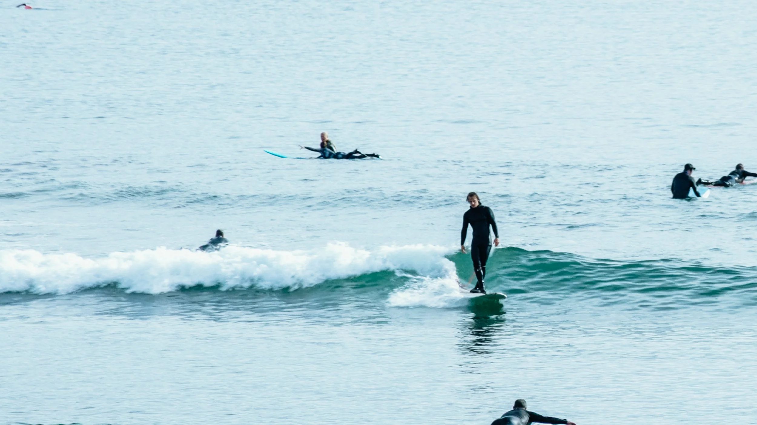 A woman surfing on a small wave at the beach, surrounded by people on surfboards and paddleboards in the water.