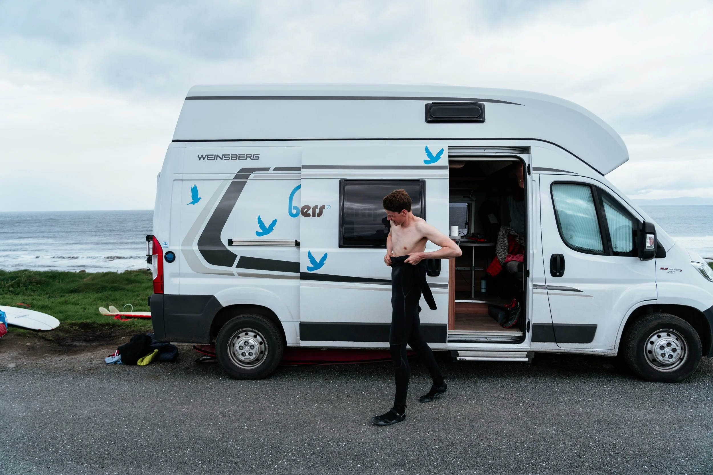 A man putting on a wetsuit next to a white camper van parked near the ocean, with surfboards on the ground nearby and the sky cloudy.