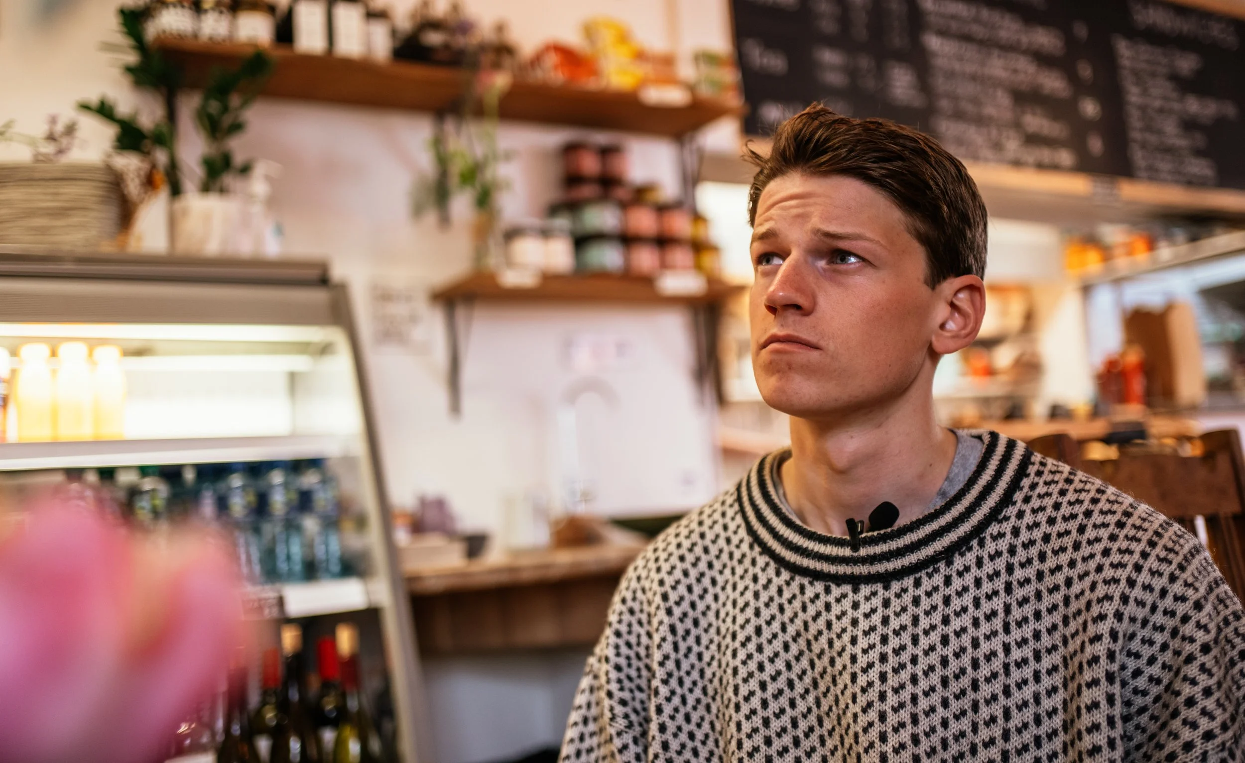 A young man with brown hair and a patterned sweater sitting inside a cafe, looking to his left with a serious expression, with shelves and a chalkboard menu in the background.
