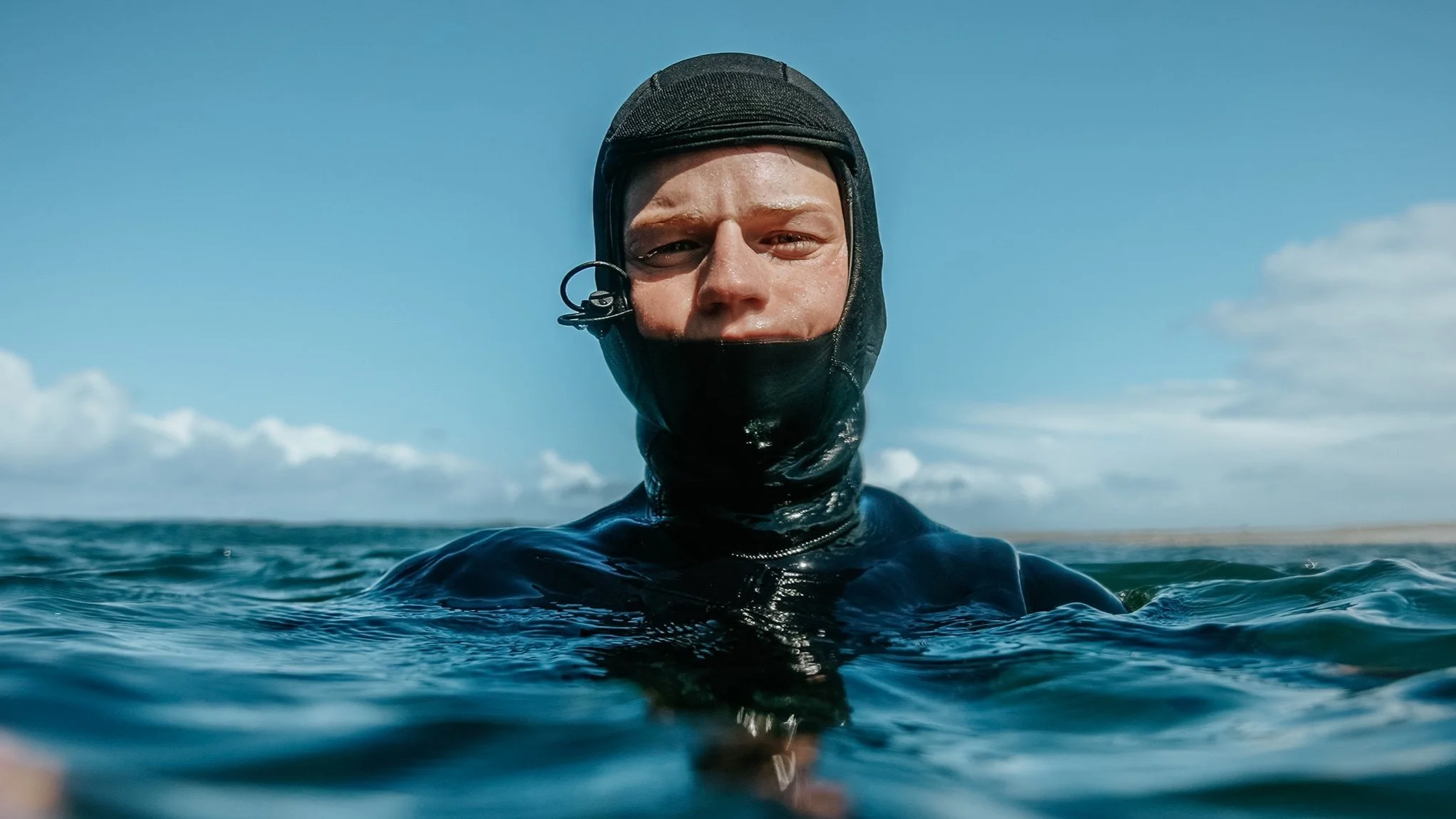 Man in a black wetsuit and cap swimming in the ocean under a partly cloudy sky.