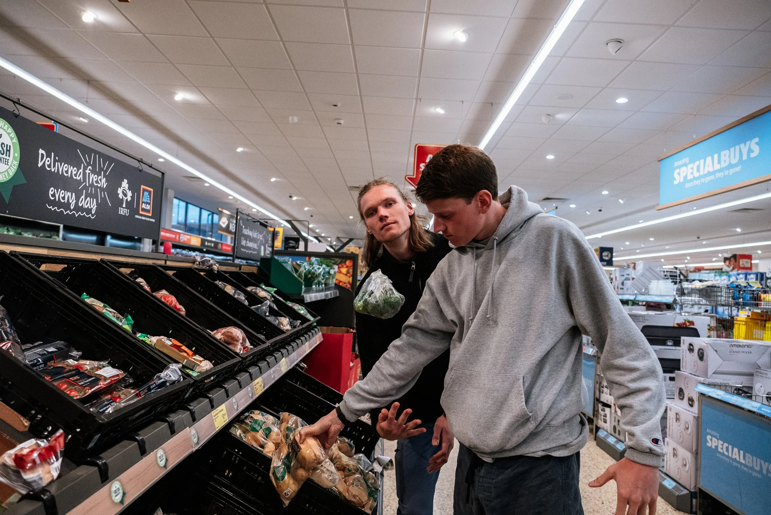 Two young men shopping for produce in a grocery store aisle, looking at potatoes, with signage for fresh produce and specials overhead.