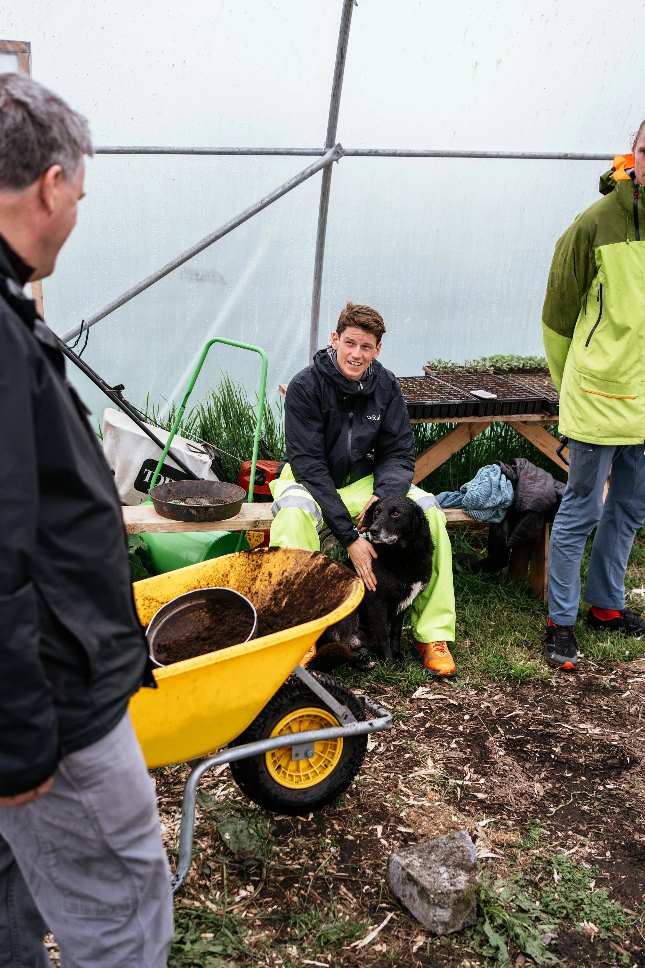 Three people inside a greenhouse with gardening supplies; one person is sitting on a bench petting a black dog, while the other two are standing nearby.