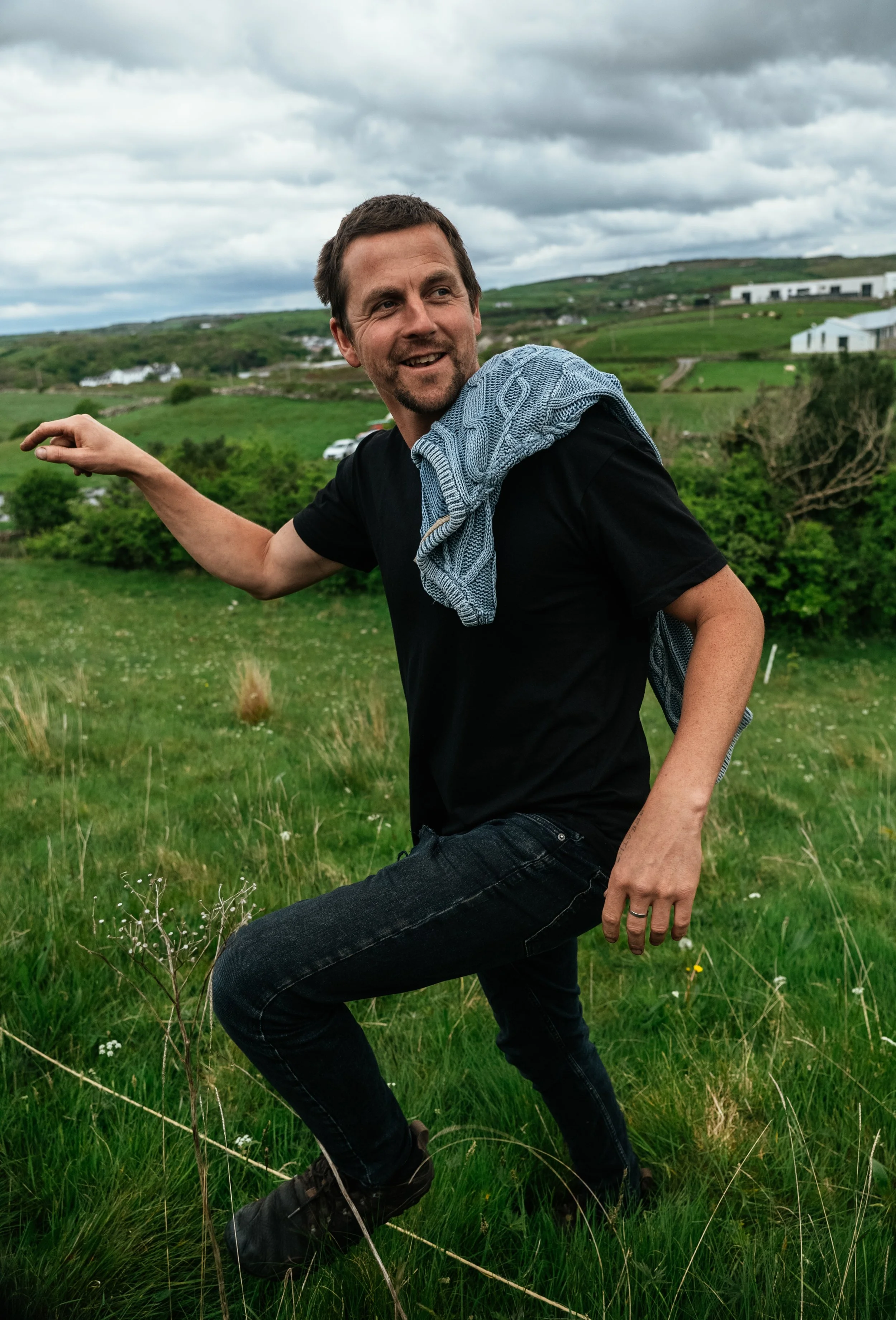 A man with a second shirt draped around his neck is outdoors in a green field, smiling and in a playful pose, with rolling hills and houses in the background under cloudy skies.