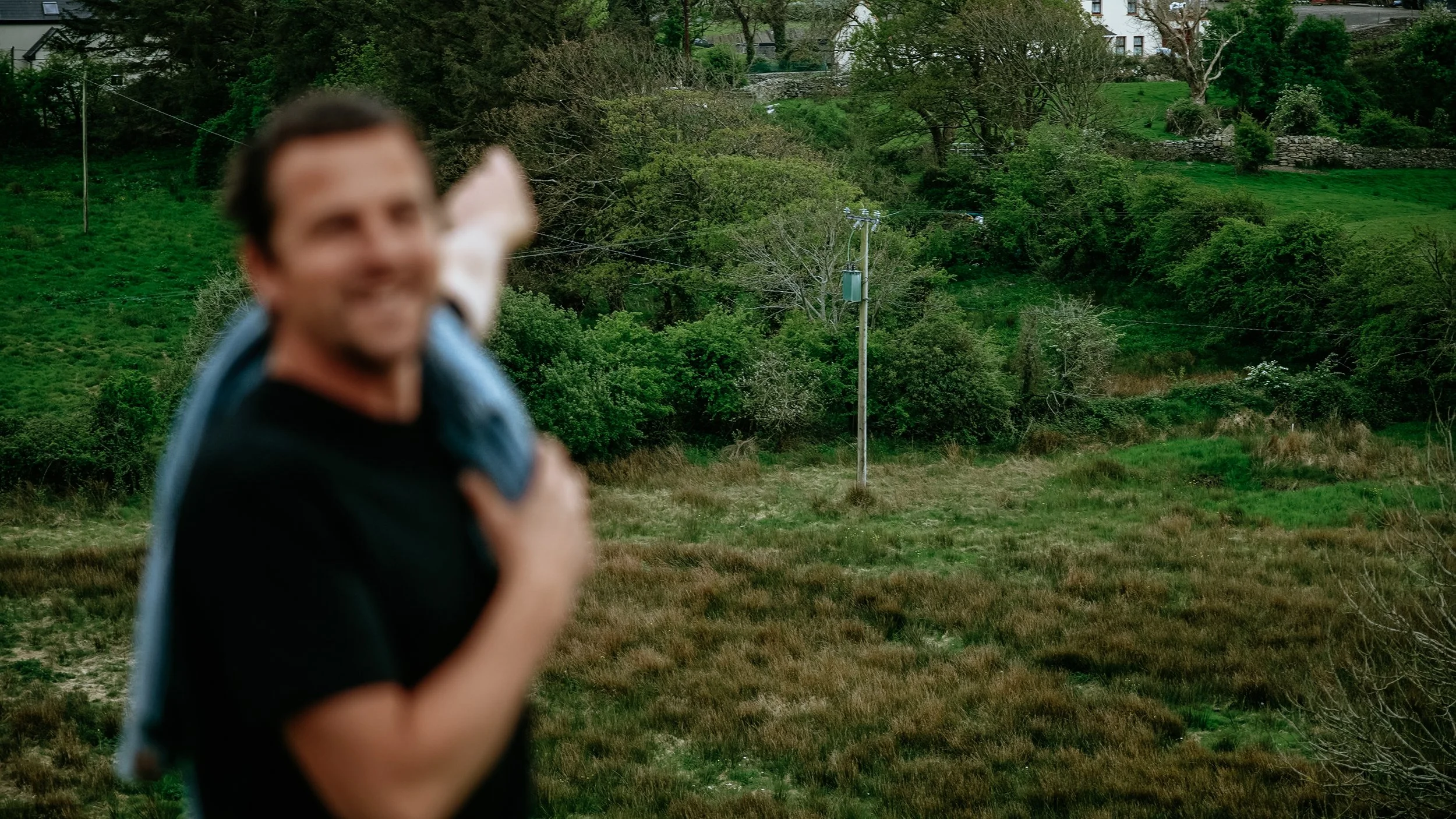 Blurred man smiling outdoors holding a jacket, with a green hilly landscape and power line in background.