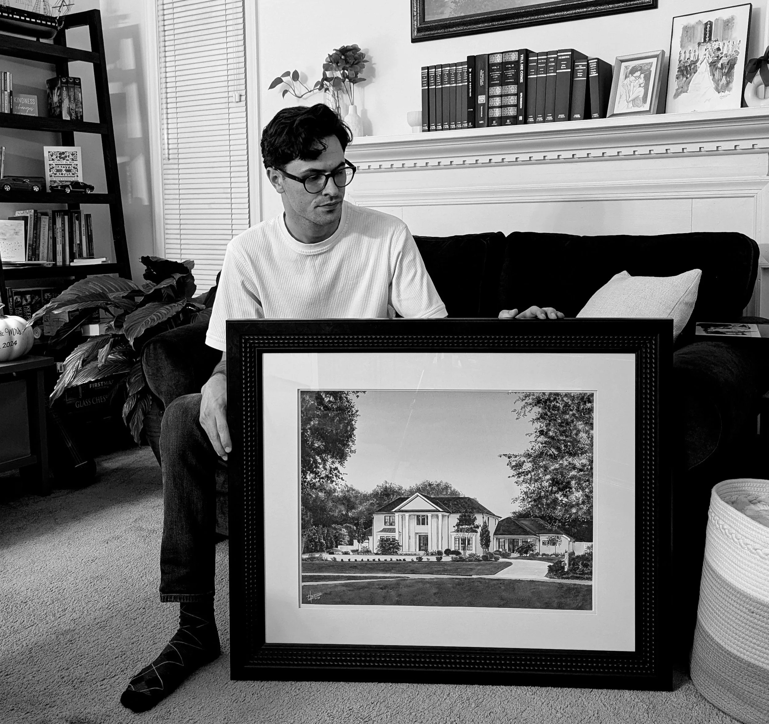 A man with dark hair, glasses, wearing a white t-shirt and dark pants, sitting on a sofa, looking at a framed photograph of a large house with a driveway and trees, placed on the floor in front of him in a living room.