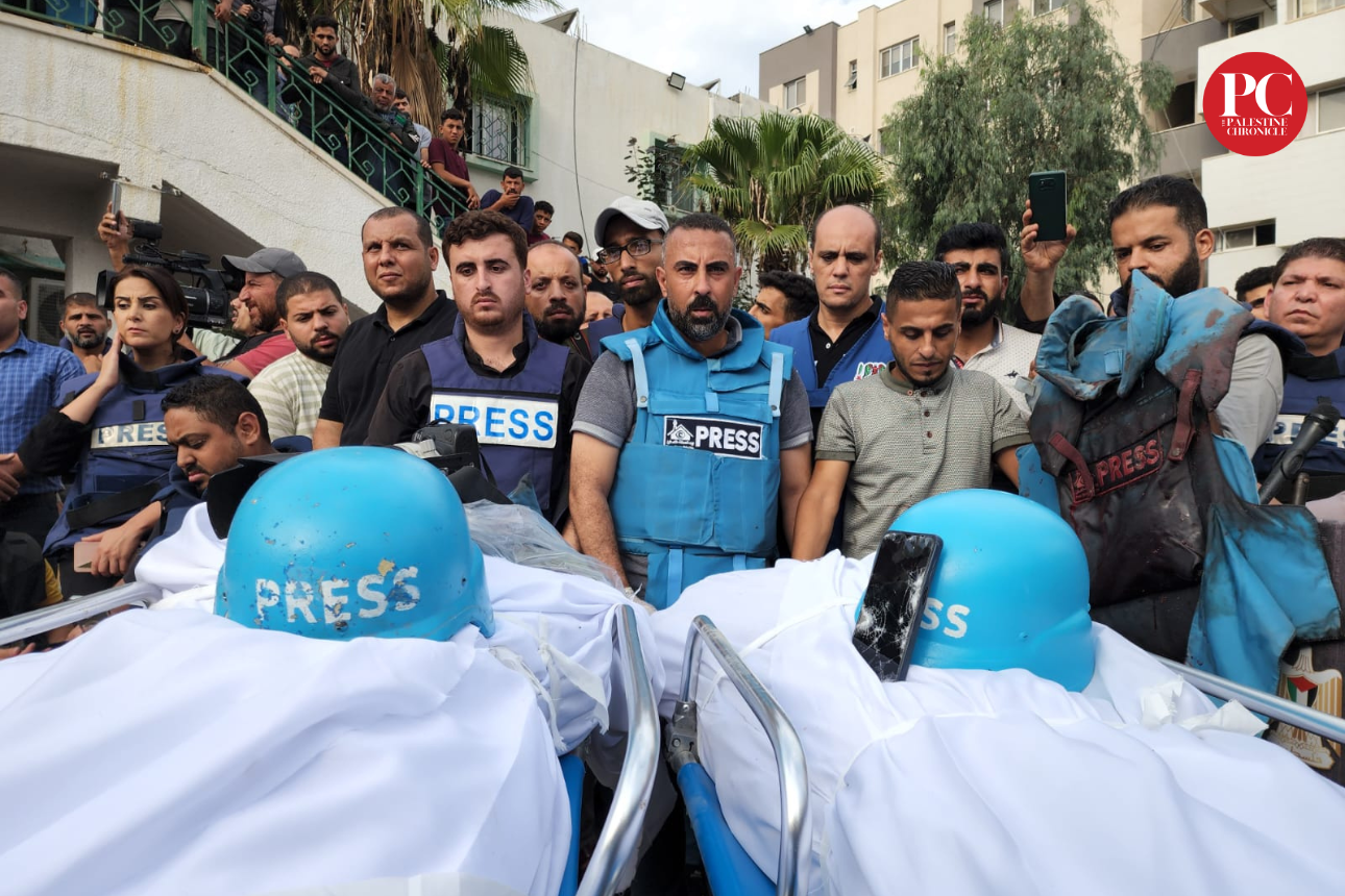 A group of journalists and bystanders gathers around a body covered with a white cloth, with two blue press helmets on top. The scene appears to be in an urban area with buildings and palm trees in the background. Some people are taking photos, and there are press vests and equipment visible.