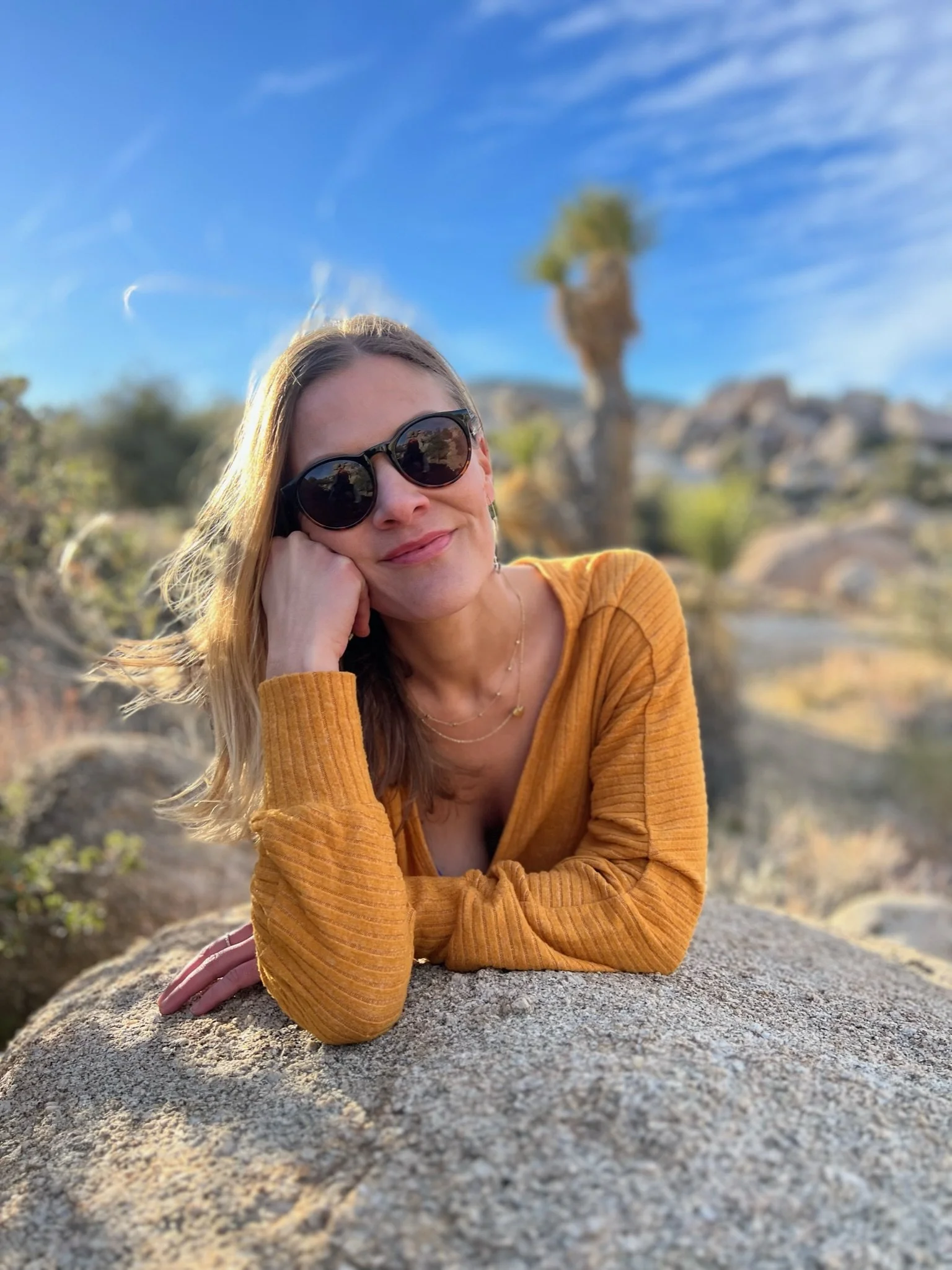 A woman wearing sunglasses and an orange top is resting her head on her hand while lying on a large rock outdoors with desert plants and a clear blue sky in the background.