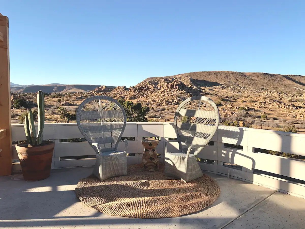 A balcony with two white wicker chairs, a small patterned side table, a potted cactus, a woven rug, and a desert landscape with rocky hills in the background.