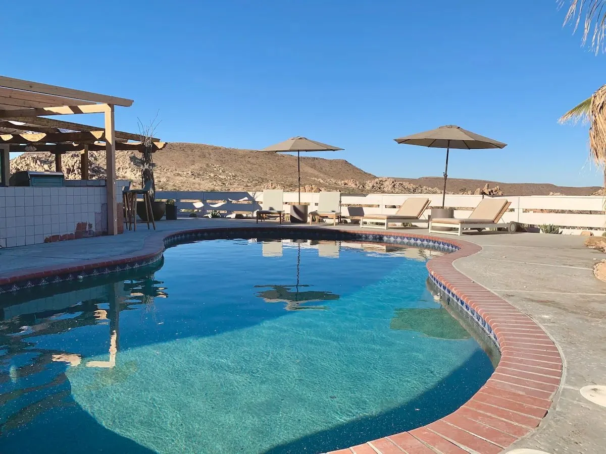 An outdoor swimming pool with a curved shape, surrounded by lounge chairs and umbrellas, set against a dry, hilly landscape under a clear blue sky.