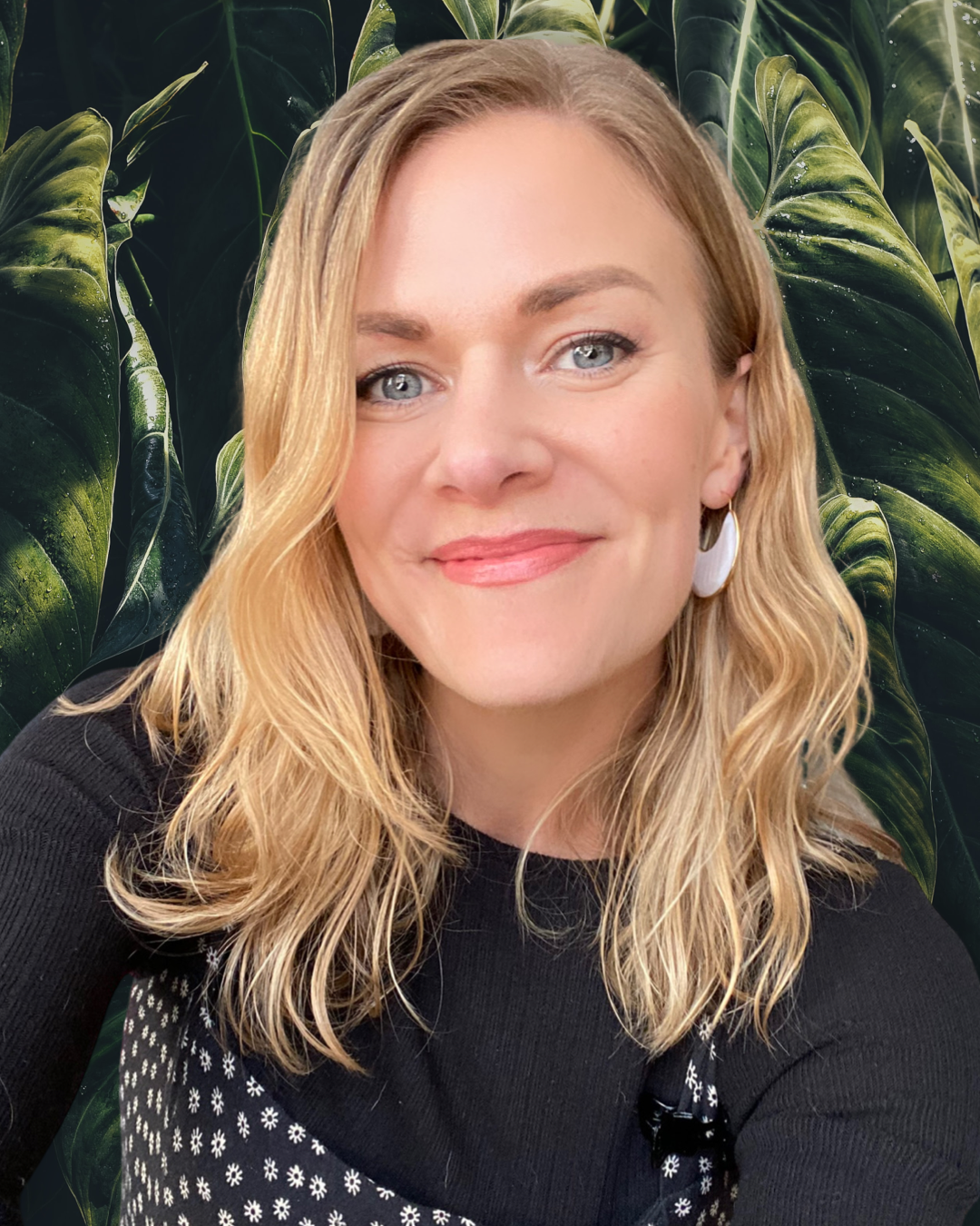 A woman with wavy blonde hair, blue eyes, and a warm smile, wearing a black top with a white floral pattern, and white earrings, standing in front of large green tropical leaves.