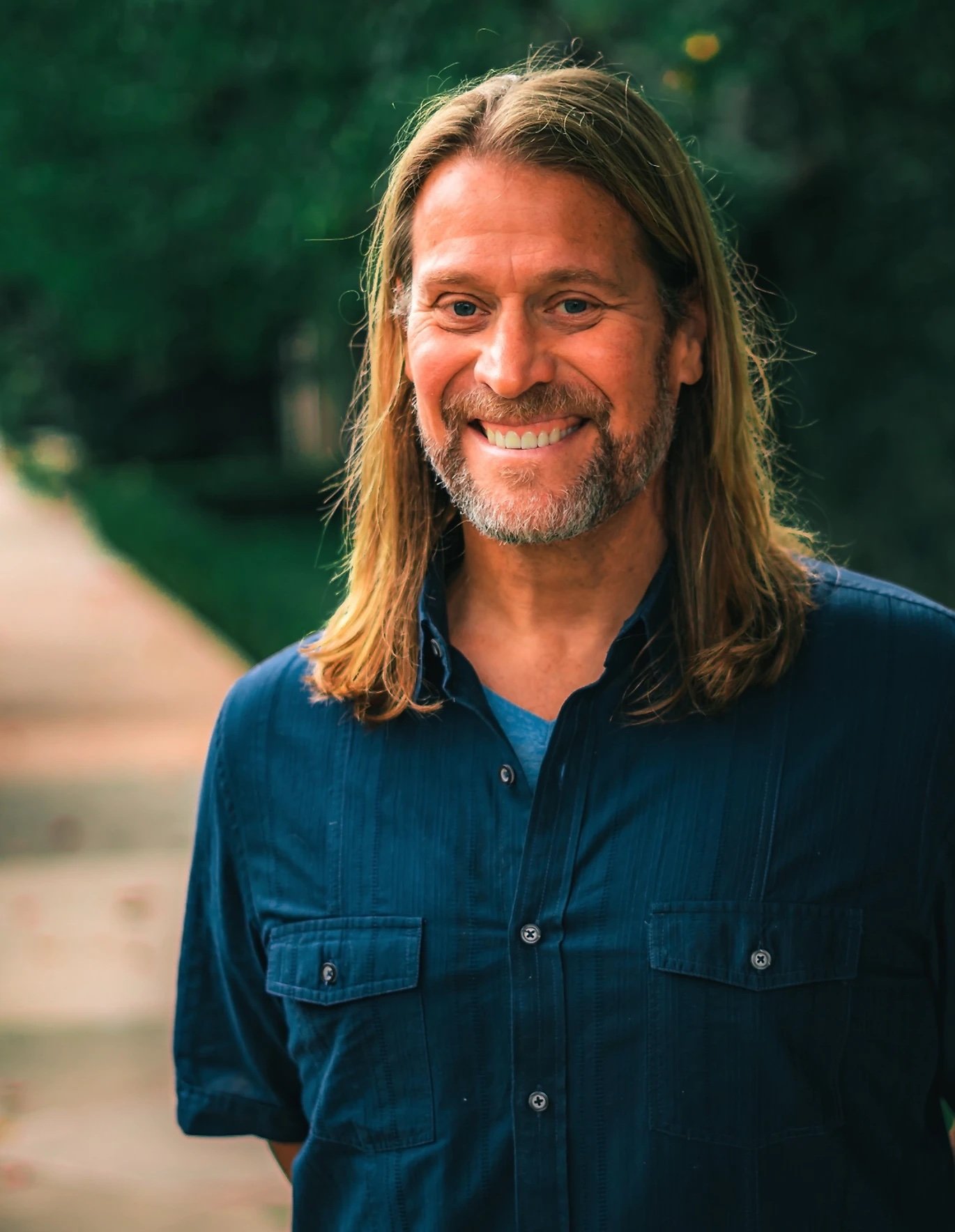 A middle-aged man with long blond hair, a beard, and blue eyes smiling outdoors, wearing a dark blue button-up shirt.