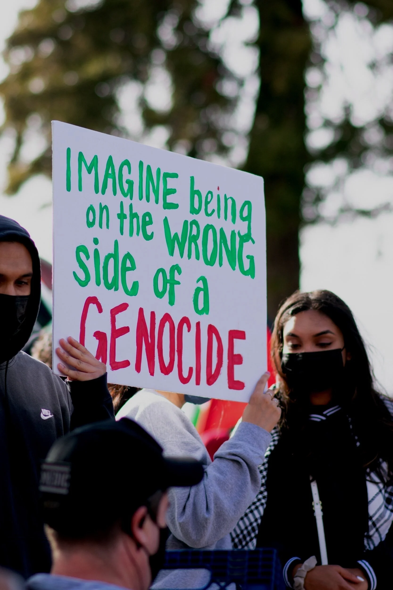 People at a protest holding a sign that says, 'Imagine being on the wrong side of a genocide,' with trees in the background.