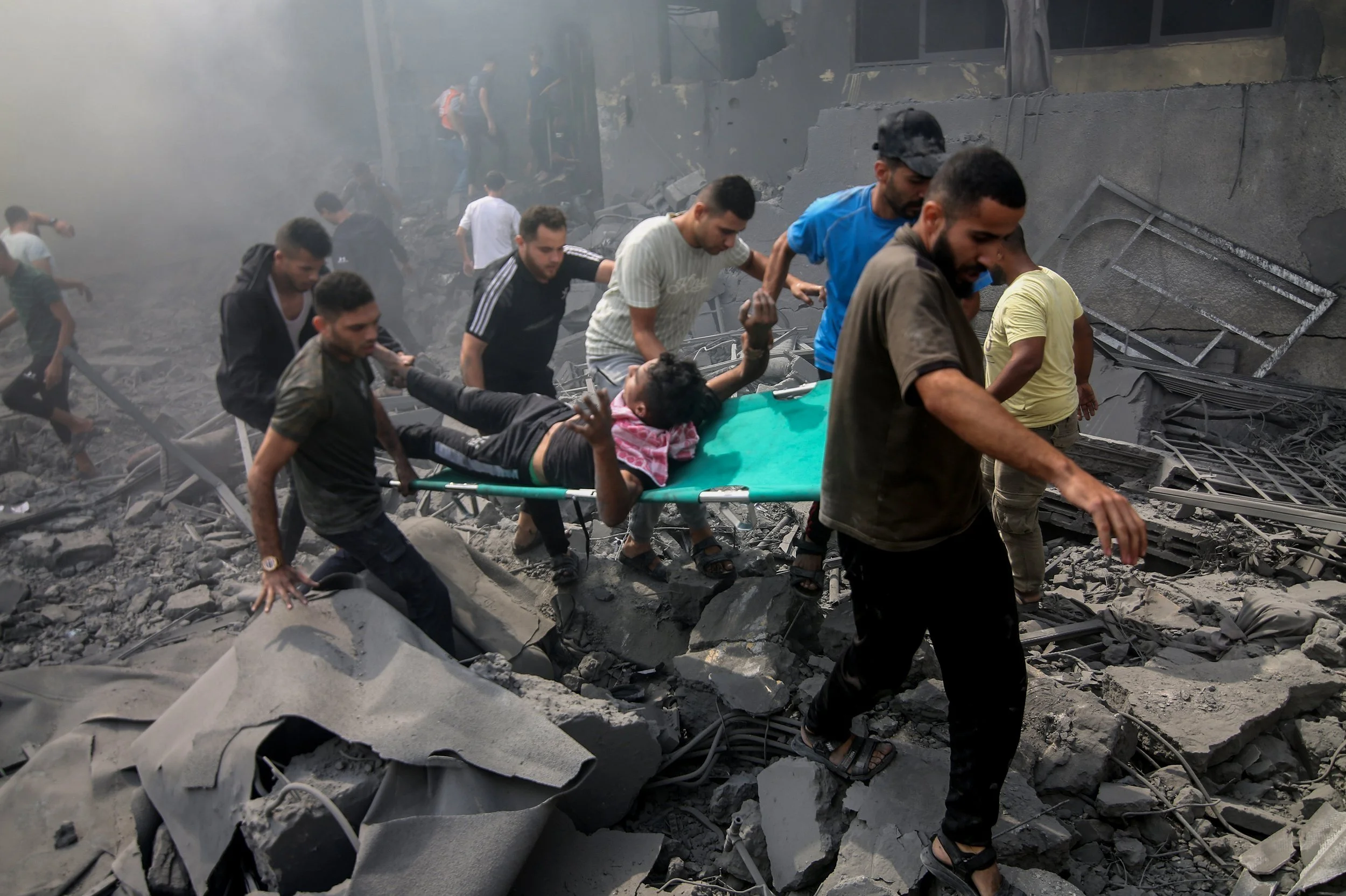 Rescue team evacuates a woman on a stretcher through rubble and debris after a building collapse, surrounded by dust and damaged structures.