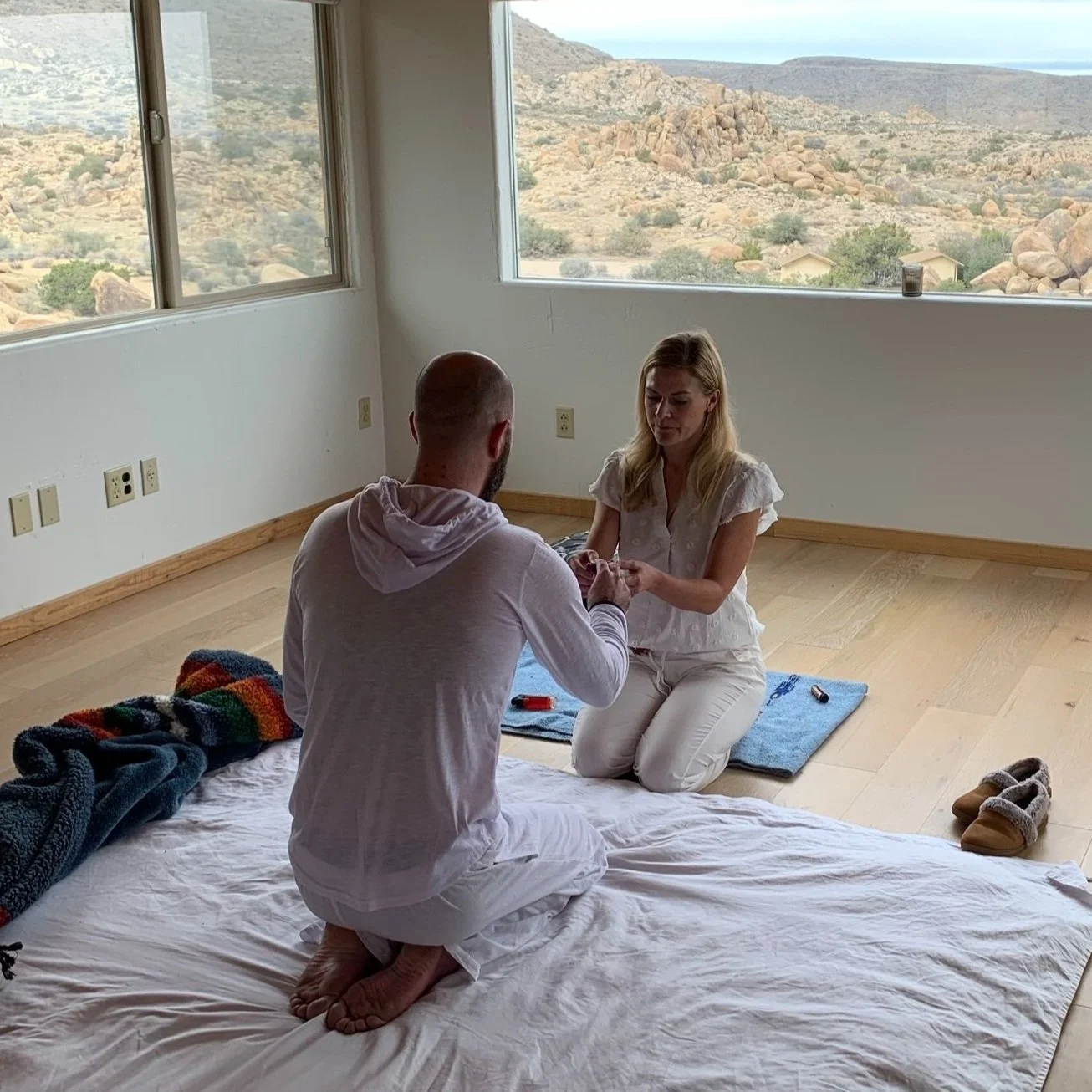 A man kneeling on a mattress and a woman sitting on the floor in a room with large windows showing a desert landscape with rocks and sparse vegetation.