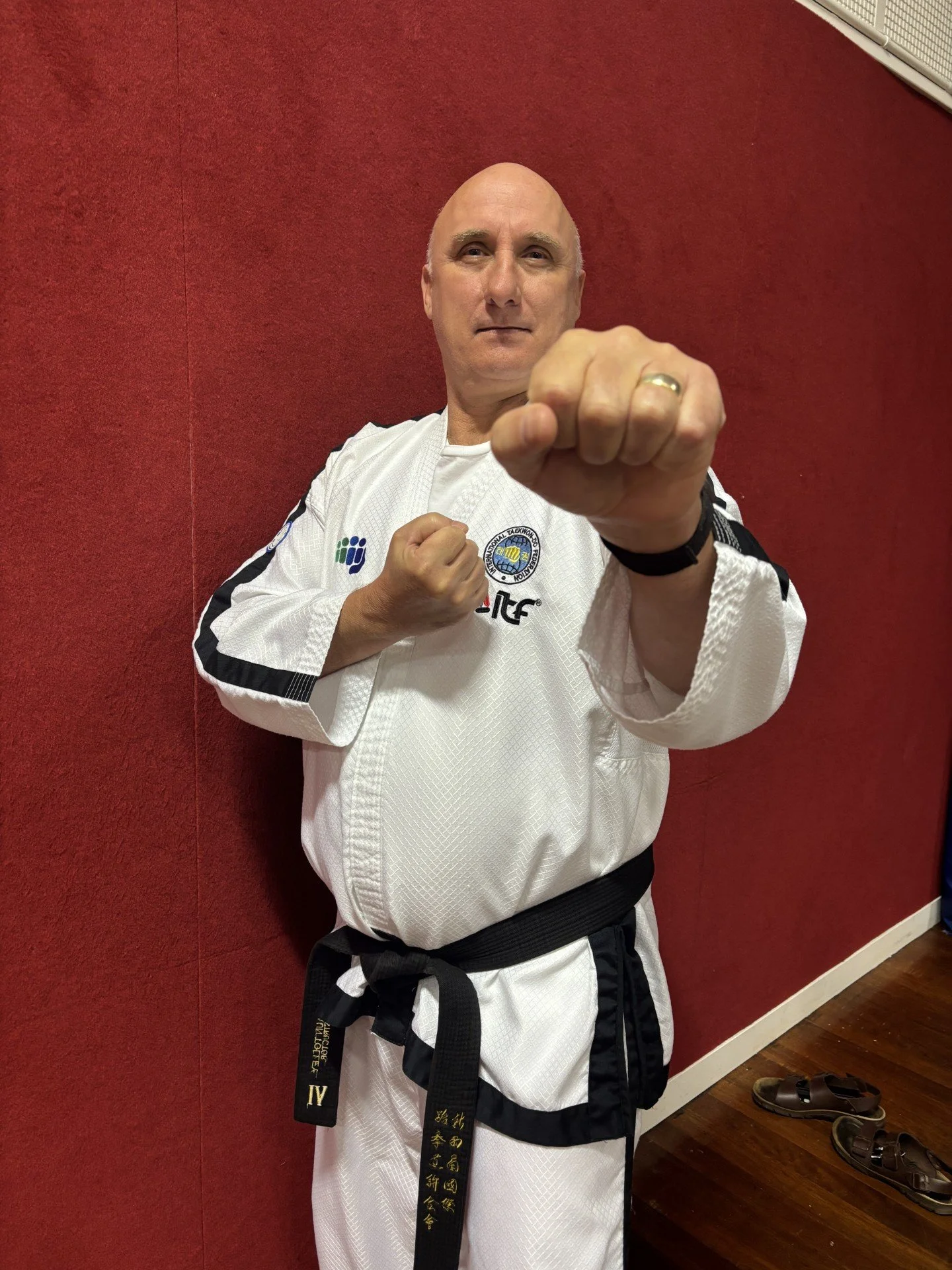 A man in a white martial arts uniform with black trim, standing in front of a red wall, making a fist with his right hand and looking confidently at the camera.