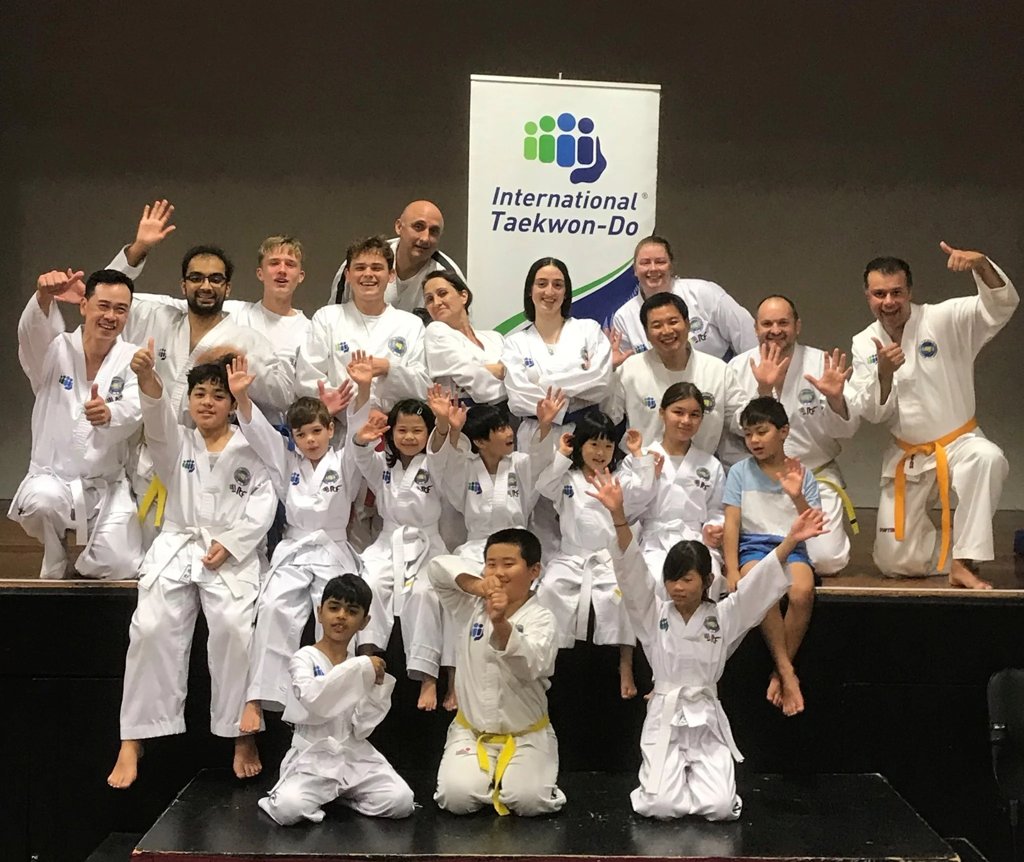 Group of martial artists, including children and adults, wearing white gis with various belt colors, posing together on a stage with a banner that reads "International Taekwondo." They are smiling, some with thumbs up, and appear to be celebrating a martial arts event.
