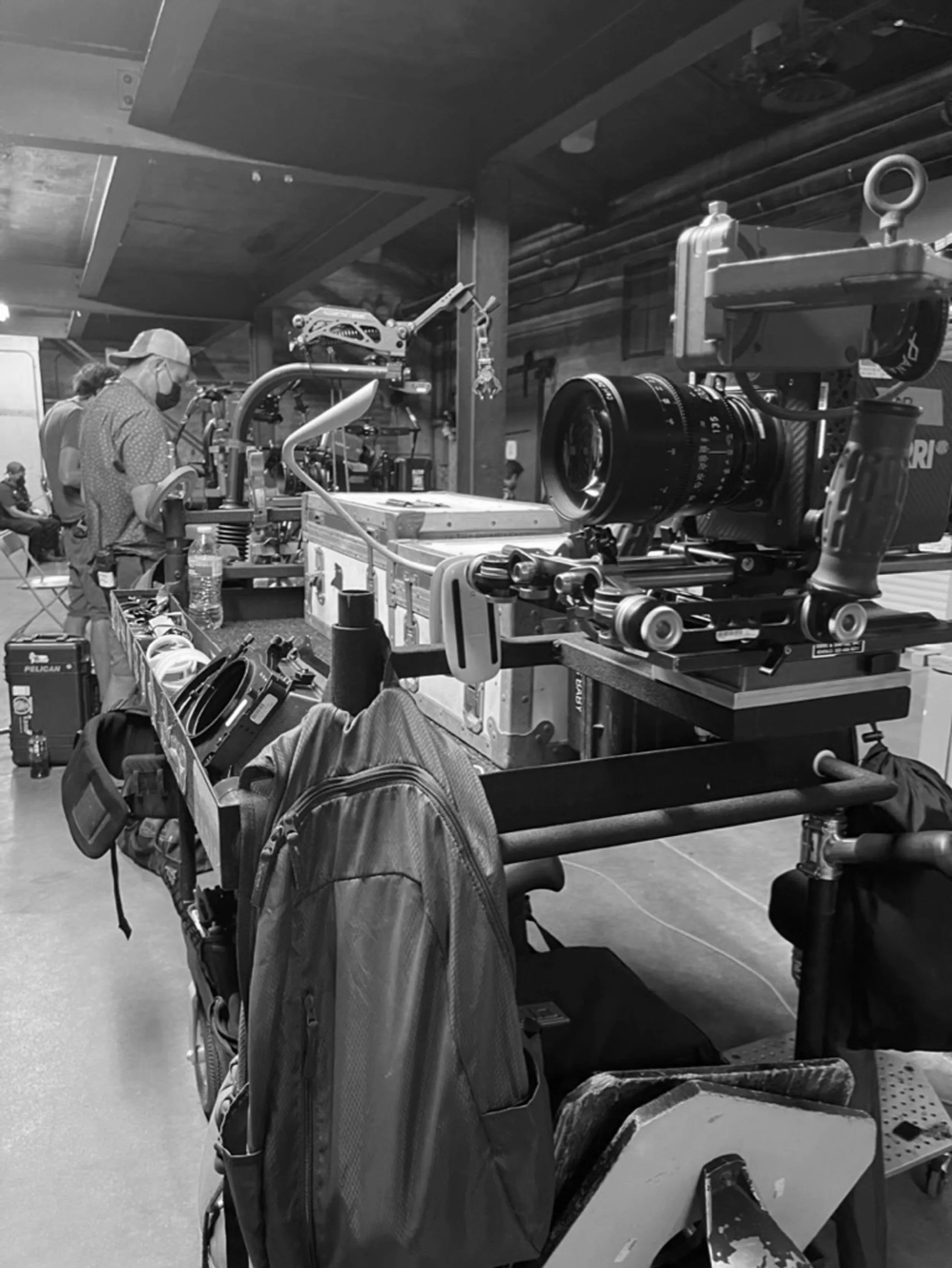 A workspace with various equipment, including a camera mounted on a dolly, a backpack, and a person wearing a mask and a hat. There are other people in the background, and the setting appears to be a studio or workshop.
