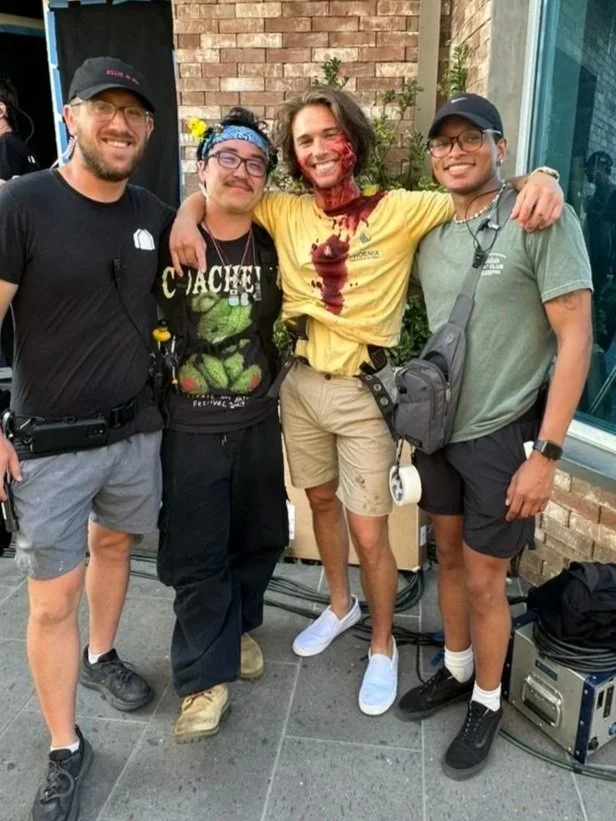 Four people stand together outdoors, smiling. The person second from the right appears to be covered in red cake or icing on face and shirt. The others are dressed casually, wearing shorts and glasses, with a brick wall and plants in the background.