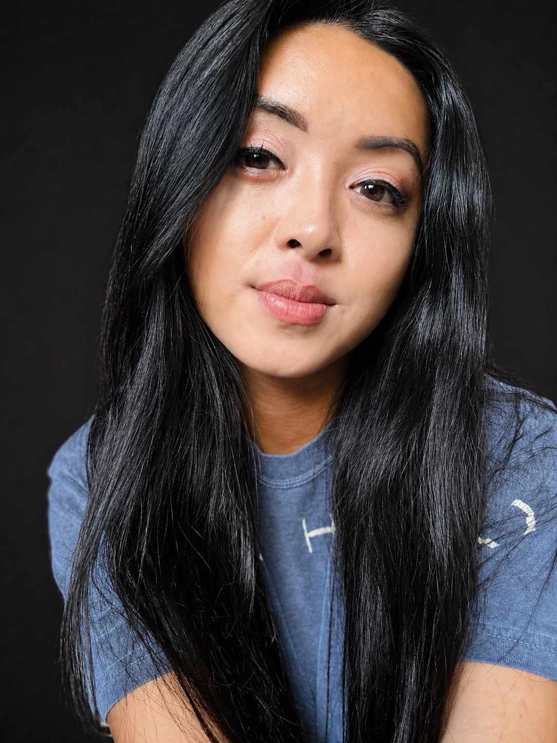 Close-up portrait of an Asian woman with long black hair, wearing a blue T-shirt, against a black background.