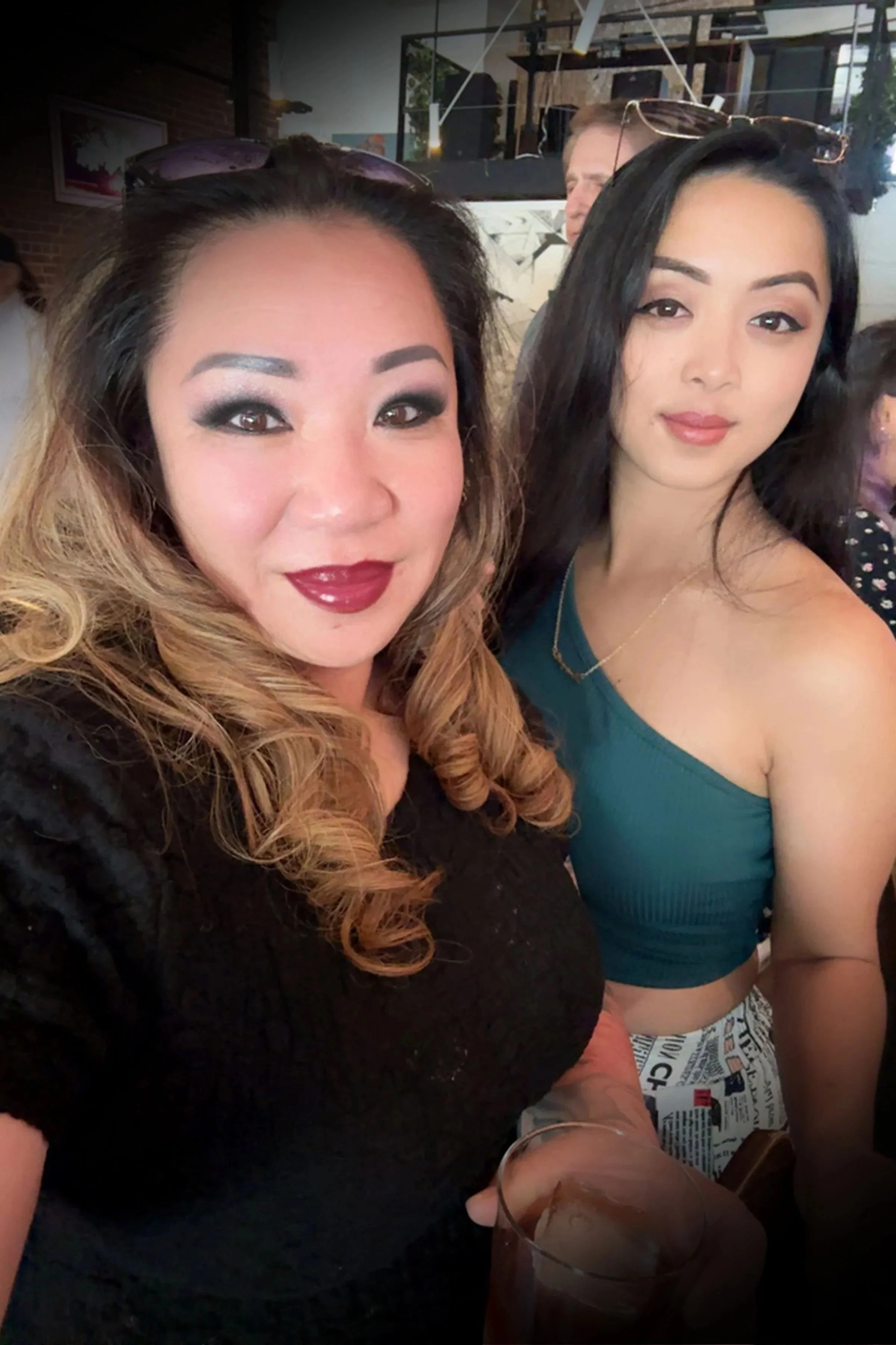 Two women taking a close-up selfie at a social gathering. The woman on the left has curly, ombre hair, makeup with dark eyeshadow, and red lipstick, and is wearing sunglasses on her head and a black top. The woman on the right has straight black hair, natural makeup, and is wearing a teal sleeveless top with a gold necklace. In the background, there are other people, a brick wall, and a wooden ceiling with lighting.