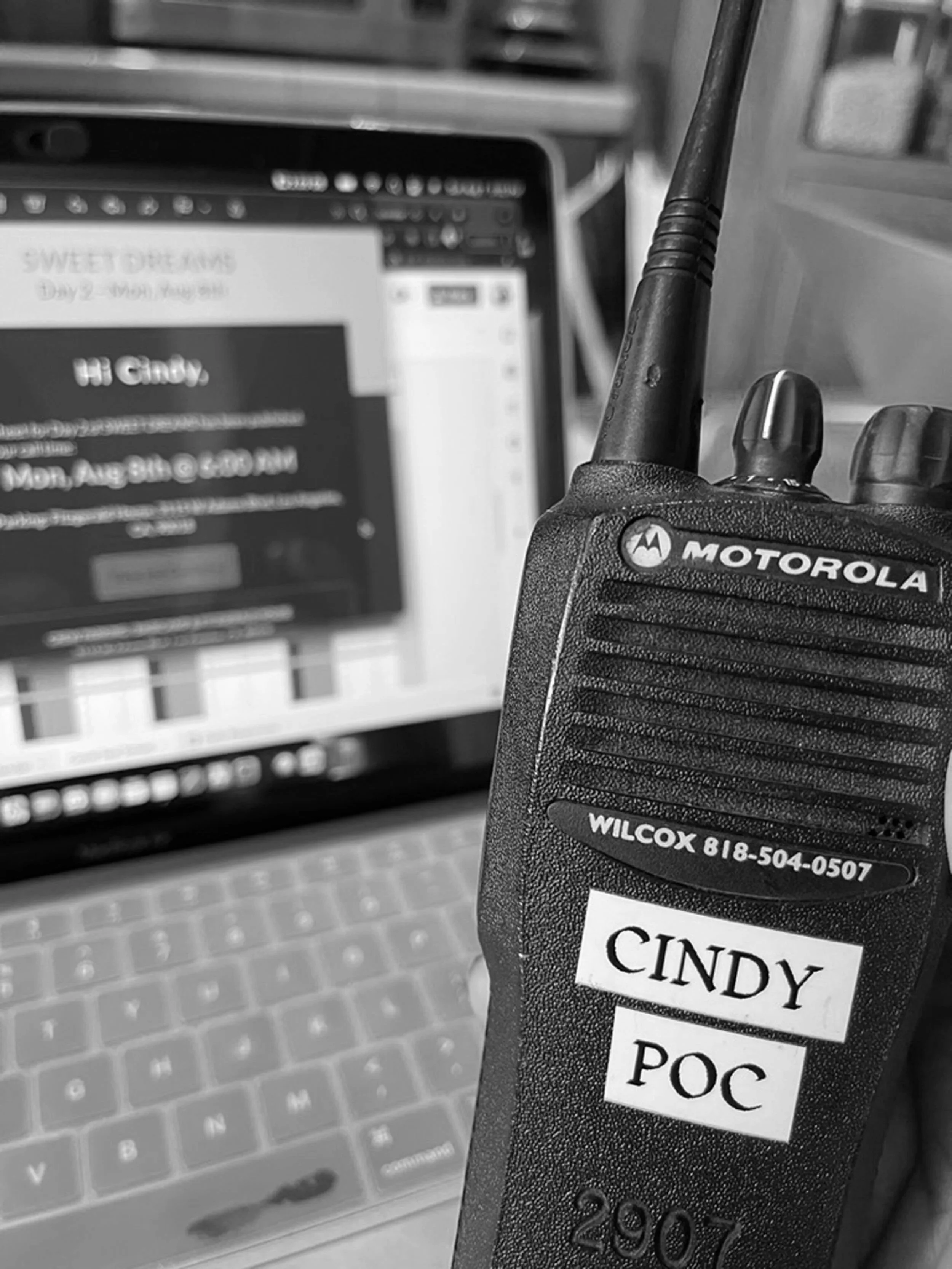 A close-up view of a Motorola walkie-talkie with a label on it reading 'CINDY POC' and a phone number, held in front of a laptop keyboard and a blurred computer screen displaying a social media webpage.