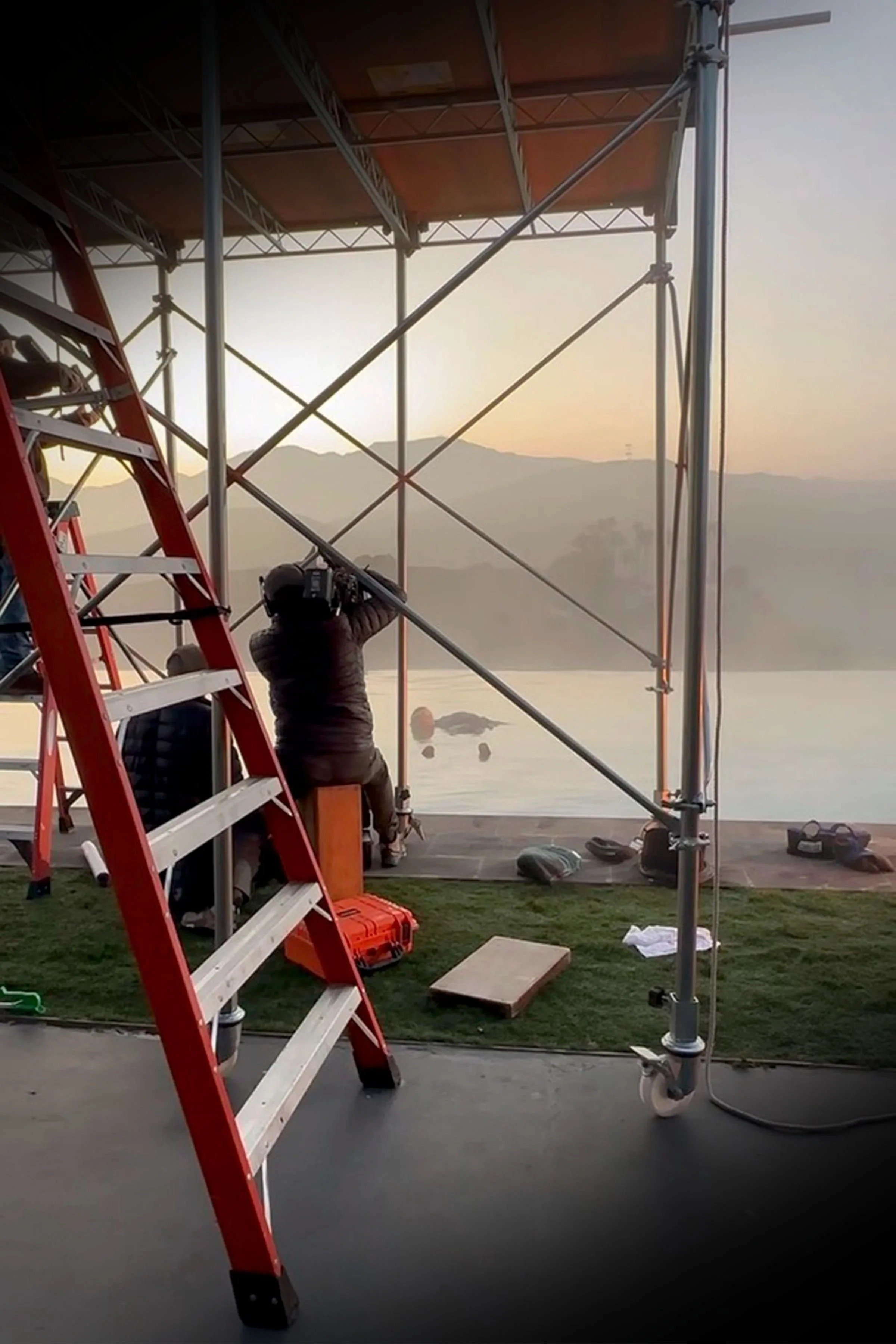 A person working on a scaffolding structure near a body of water during sunset. Ladders and equipment are visible, with mountains in the background.