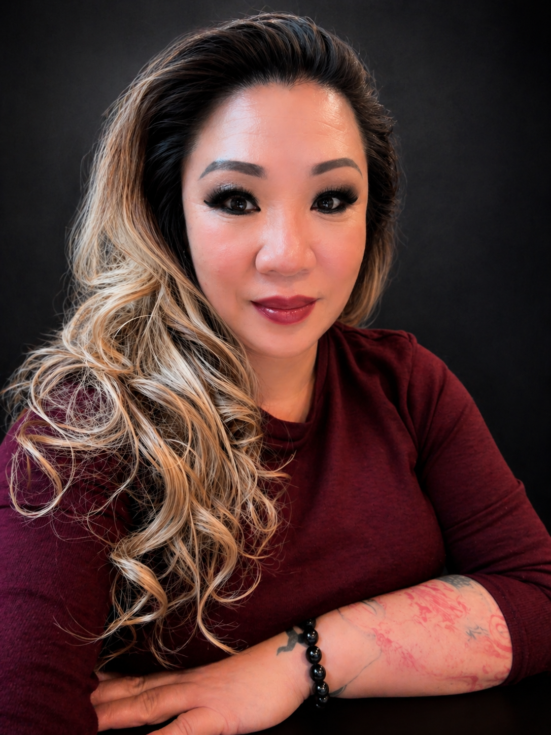 A woman with long, curly blonde and dark hair, wearing a maroon long-sleeve shirt, sitting with her arms crossed, looking into the camera with a black background.