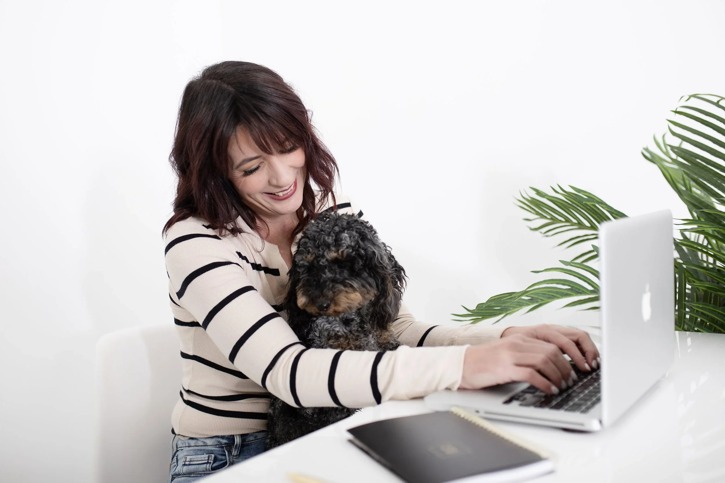 A woman with shoulder-length dark hair is smiling as she holds a black and brown curly-haired dog in her arms while working on a computer at a white desk. There is a green leafy plant on the right side of the image.