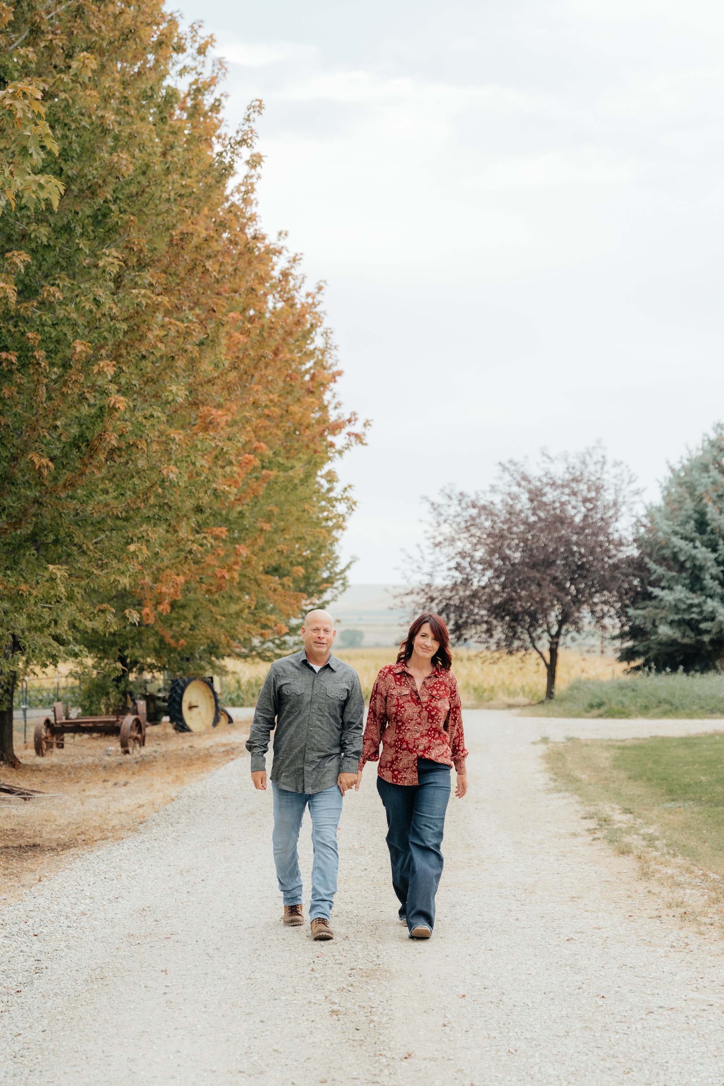 A man and woman walking hand in hand on a gravel path near trees with autumn leaves, farm equipment visible in the background.